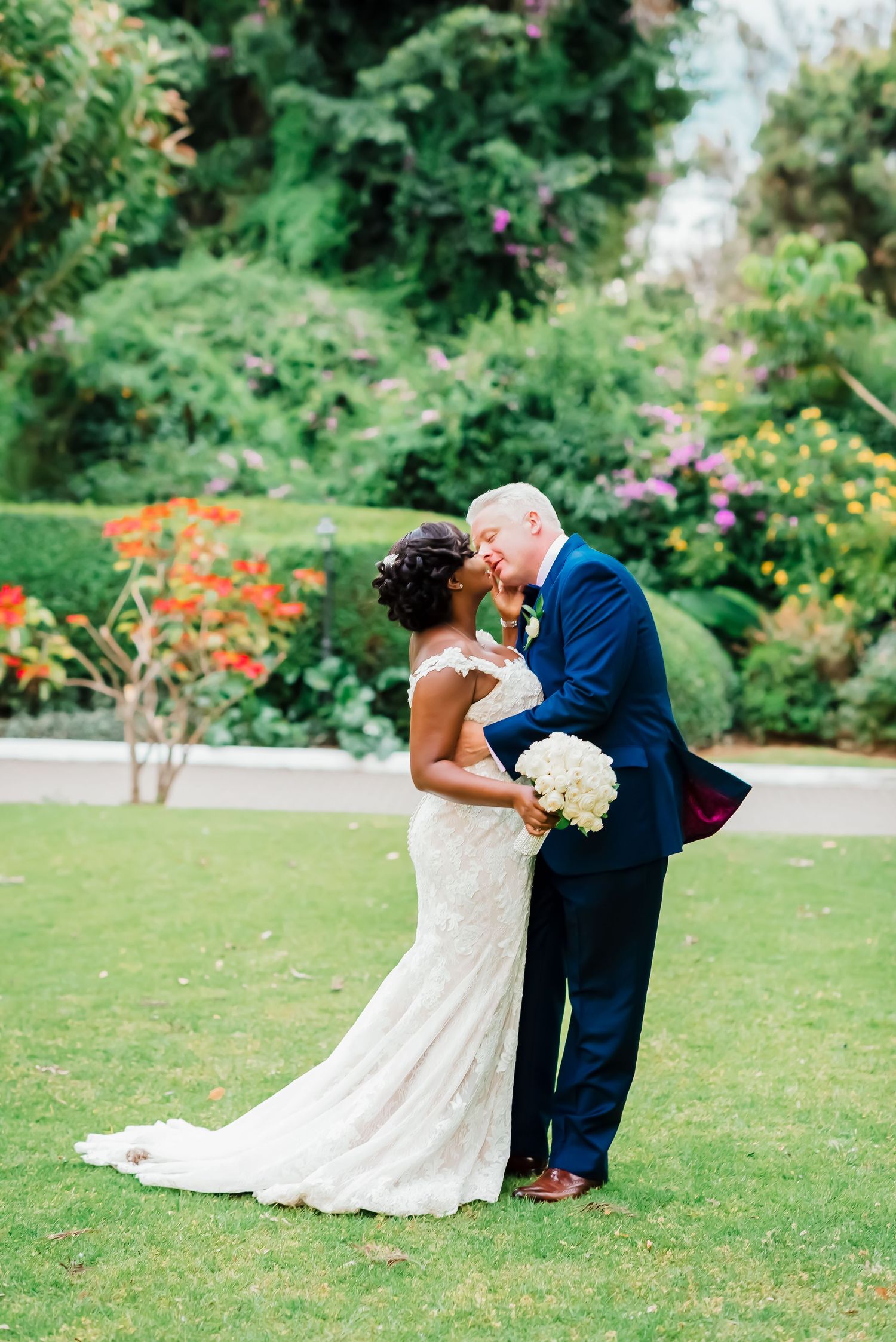 A romantic wedding couple shares a tender kiss on a lush green lawn surrounded by colorful garden flowers.