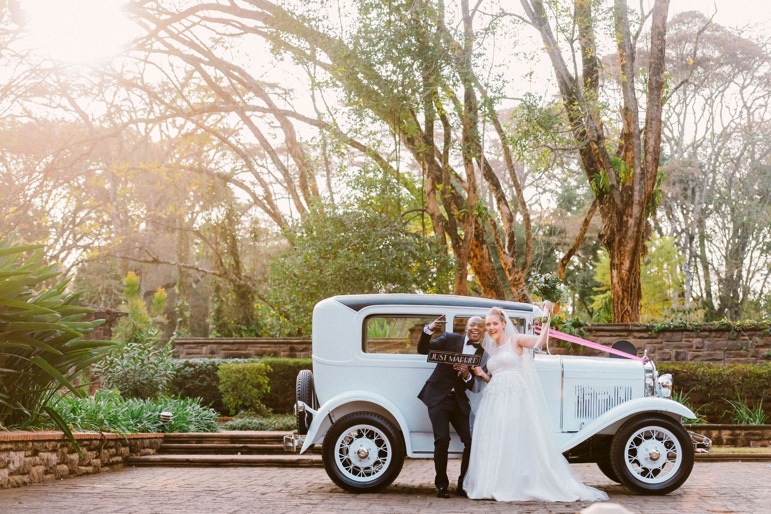 A couple poses in front of a classic white vintage car in a park setting during sunset.