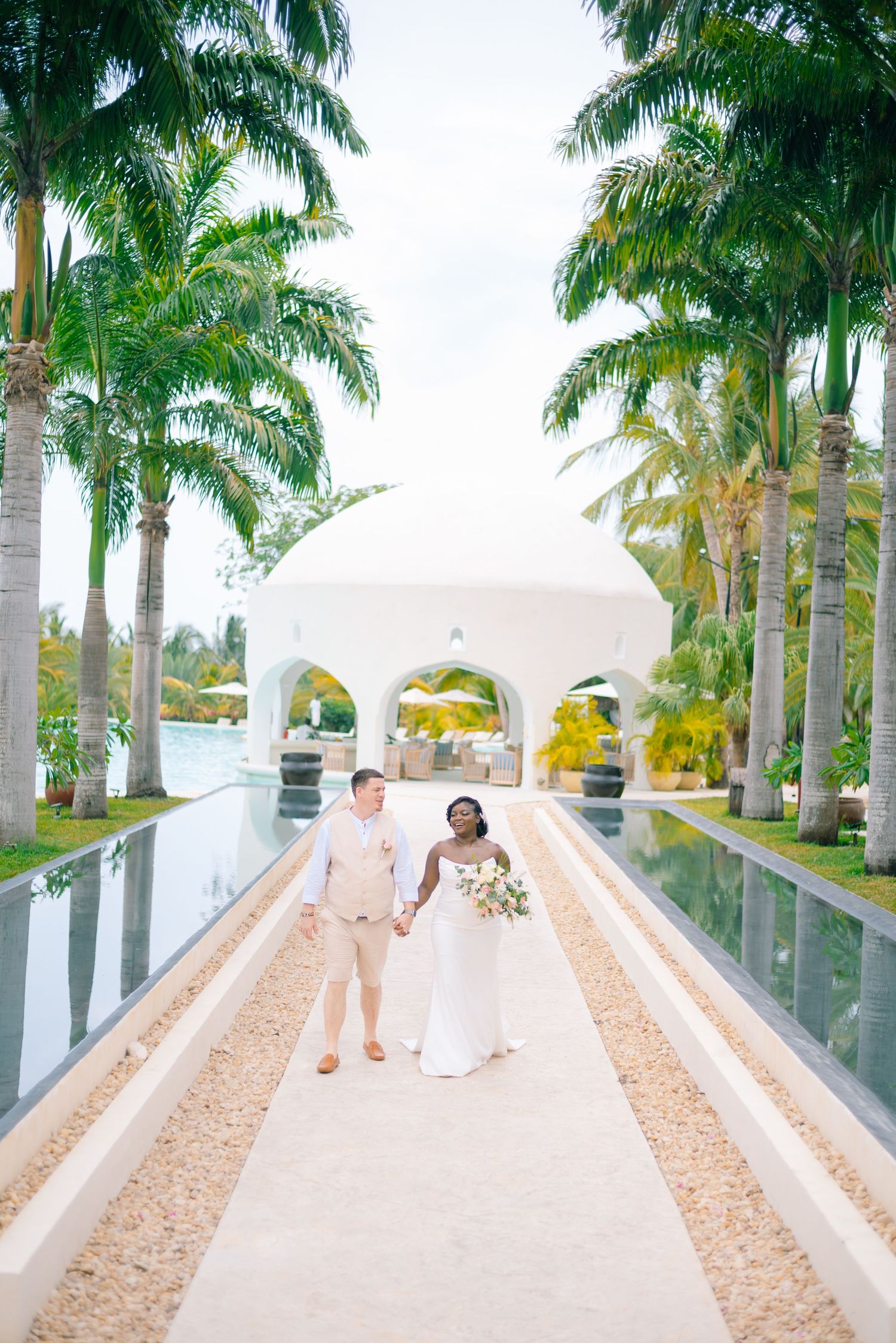 A palm tree-lined walkway with white arches leads to a tropical beachfront wedding ceremony setup.