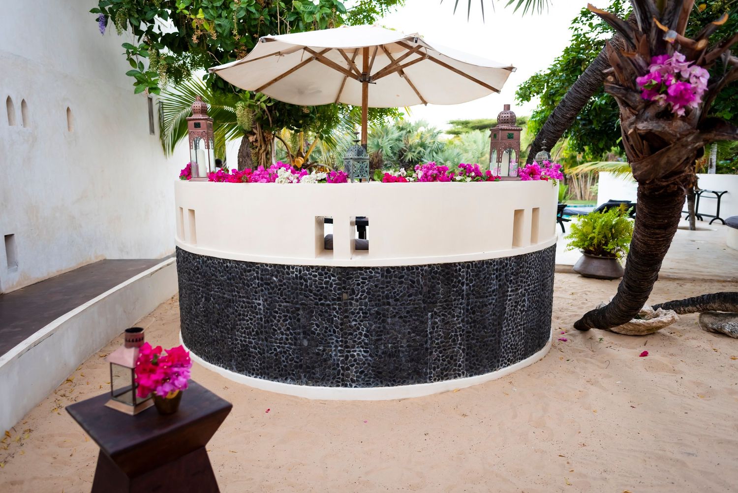 A tranquil garden courtyard features a white umbrella, pink flowers, and lush greenery around a stone pathway.