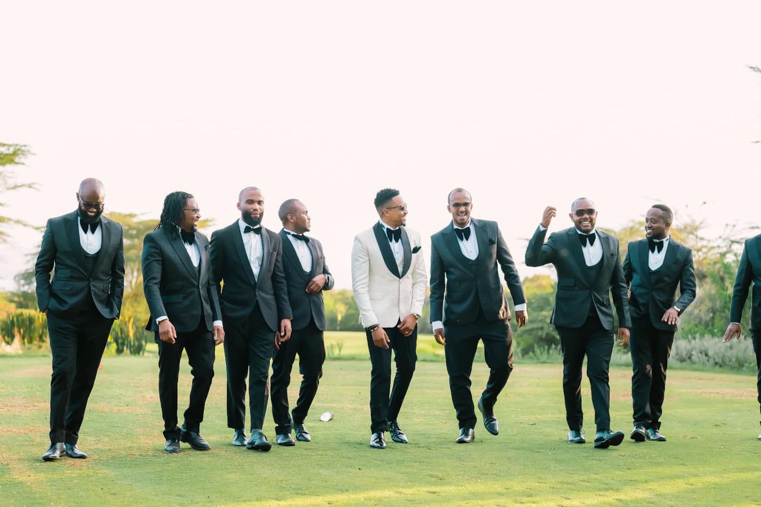 Wedding party in black tuxedos jumps in sync on a grassy lawn during outdoor photo session.