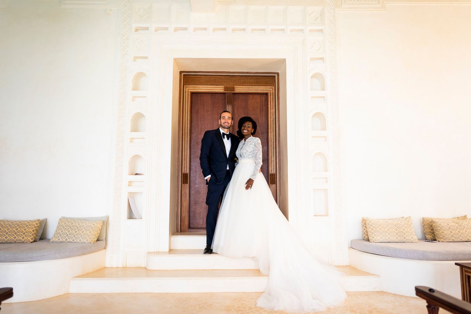 A couple in formal wedding attire stands in a doorway of a light-colored Mediterranean style building.