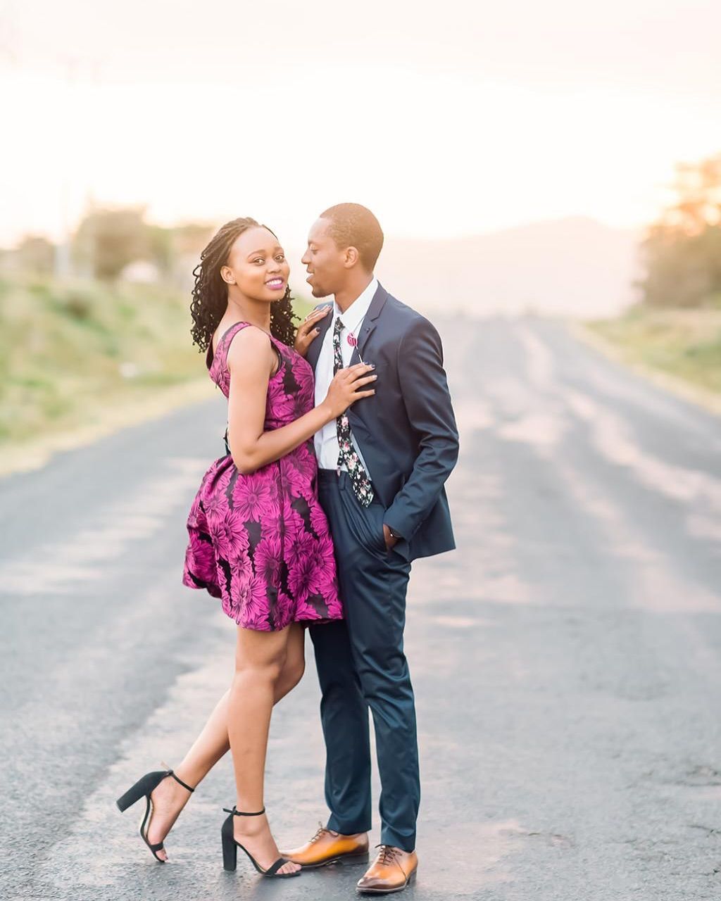 A couple in formal attire share a romantic moment on an empty road at sunset, with mountains in the background.