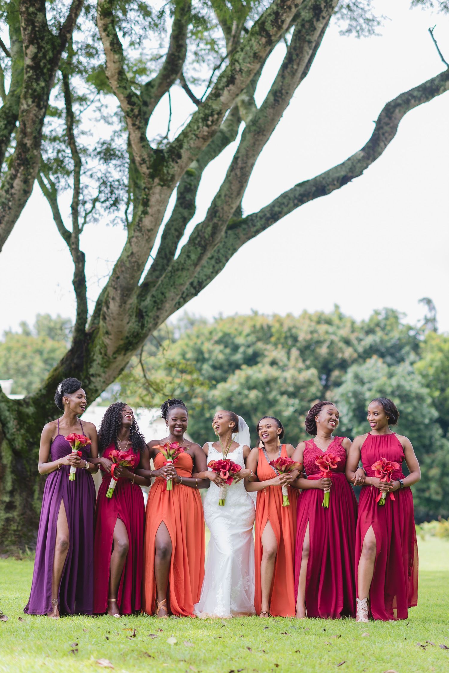A wedding party wearing vibrant shades of burgundy, purple, and coral dresses poses together outdoors on a sunny day.