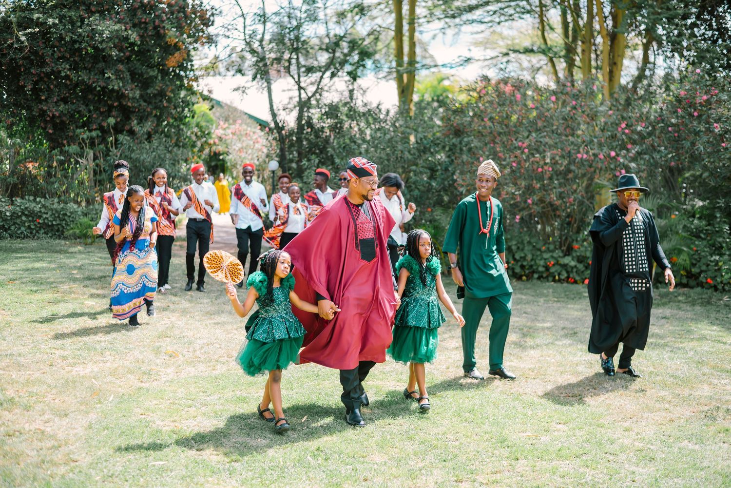 A group wearing graduation robes and formal attire walk together on a grassy path during a celebration ceremony.