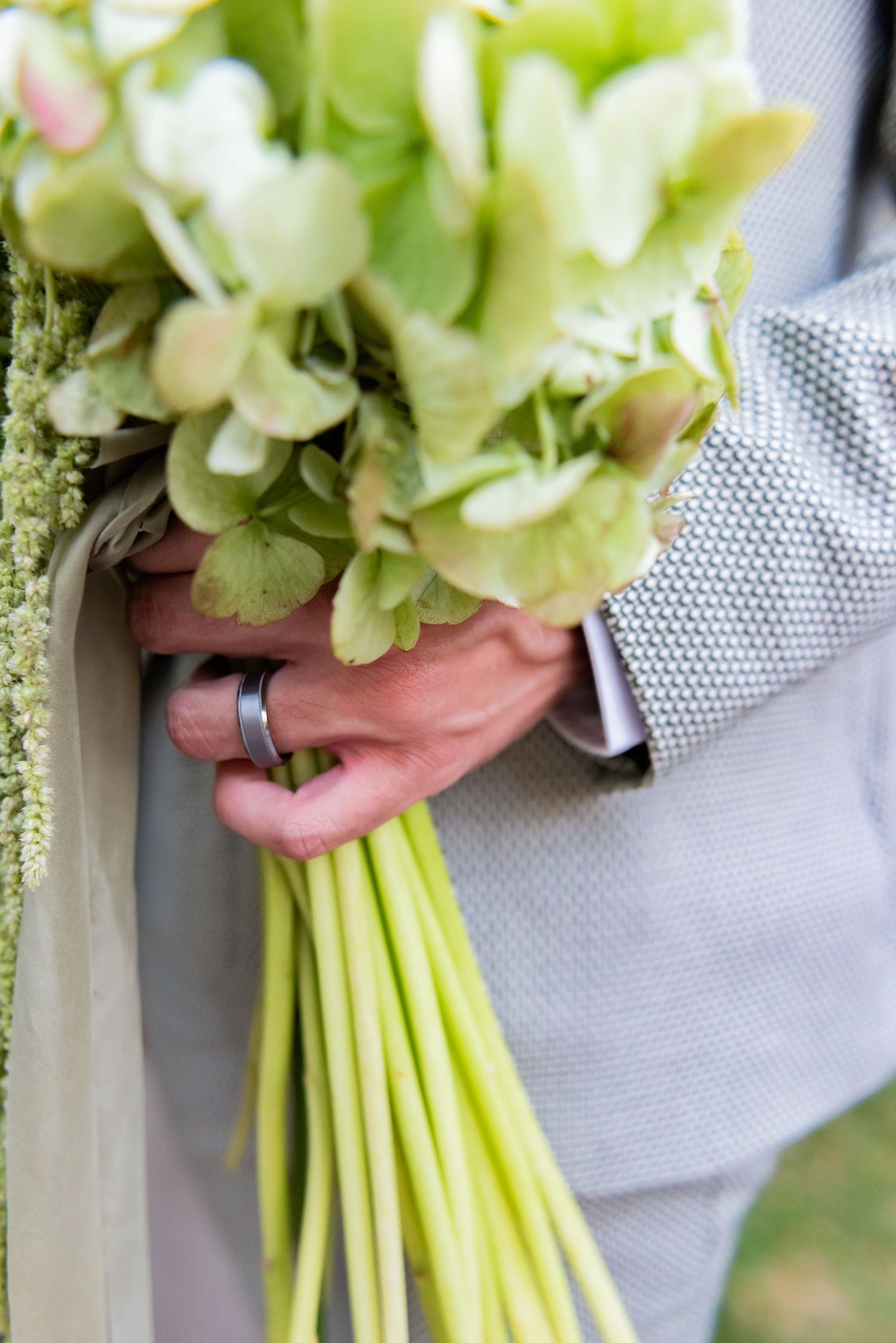 Close-up of hands holding a green and white wedding bouquet with a wedding ring visible against a light gray suit.