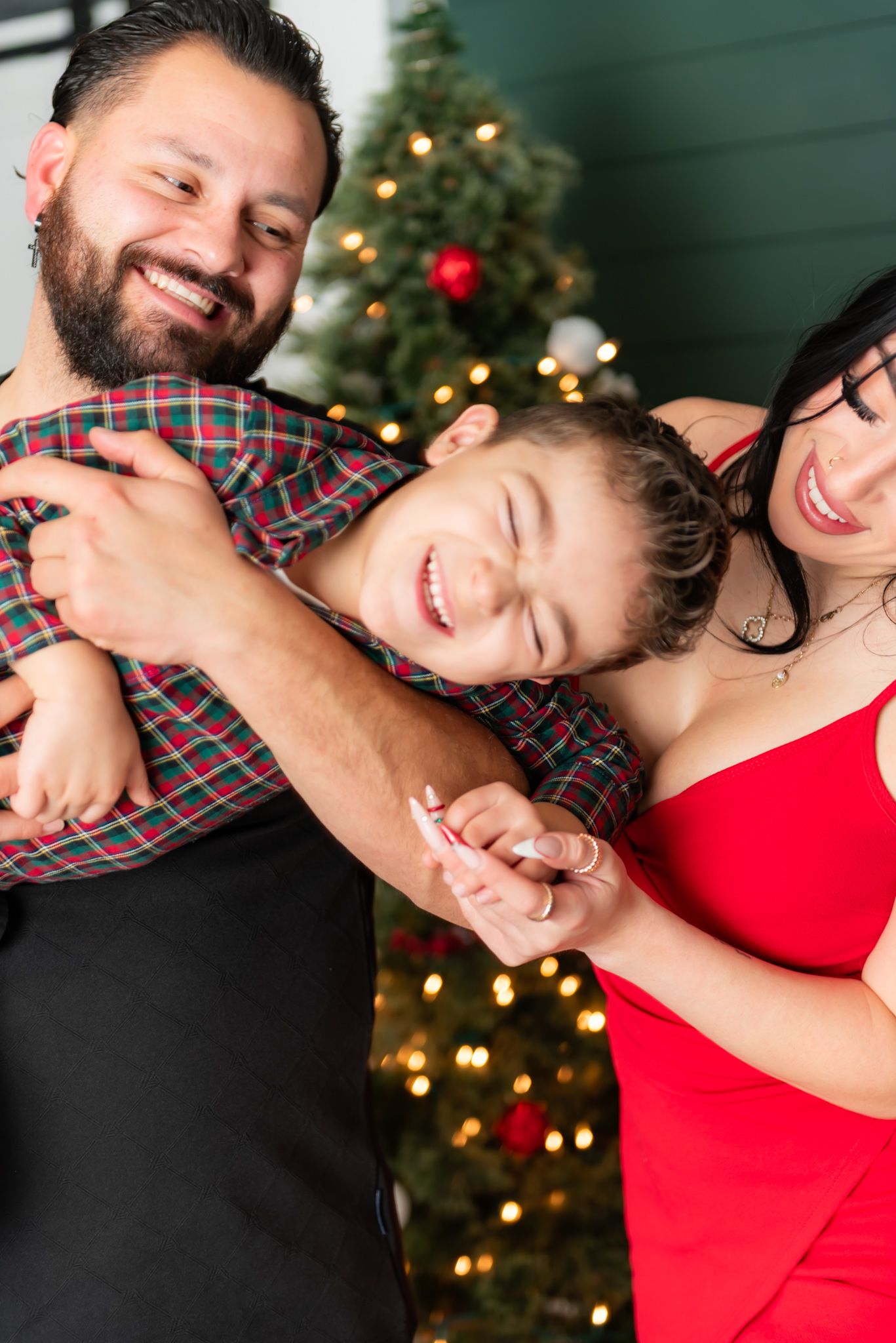 Joyful couple in red and plaid embrace and laugh together in front of a Christmas tree with twinkling lights.