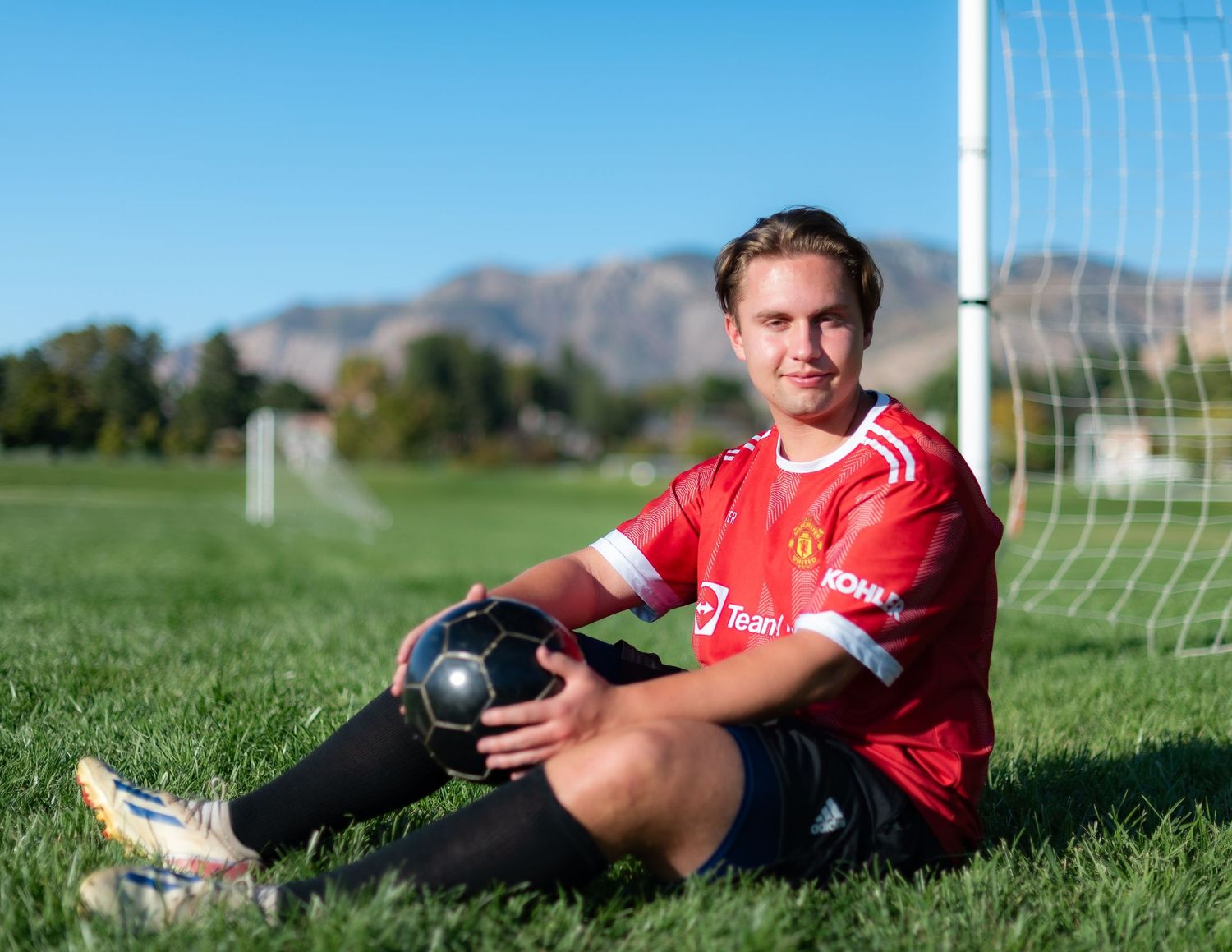 Soccer player in red jersey sitting by goal post on green field.