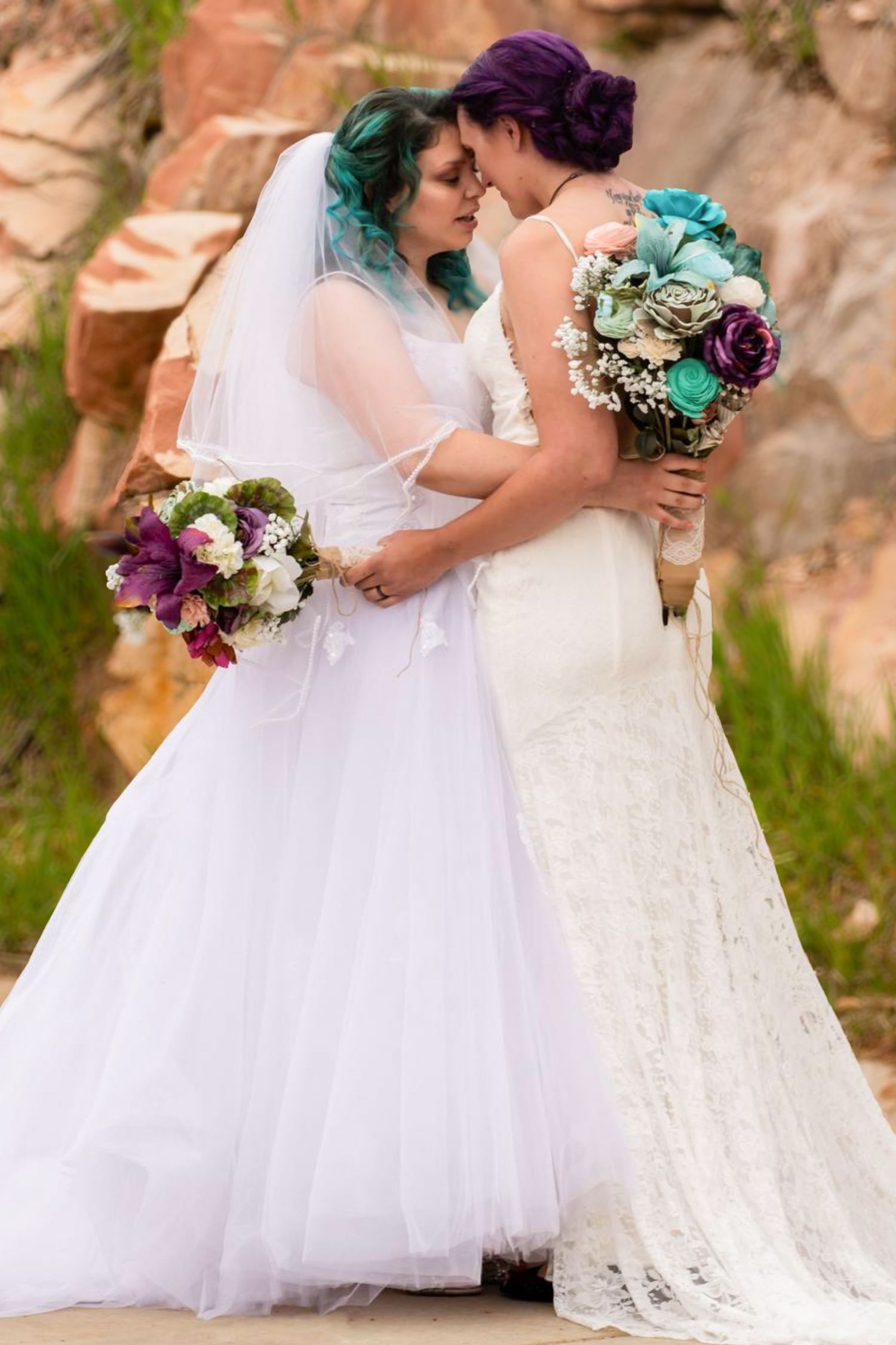 Two brides in white wedding dresses embracing with colorful bouquets against red rock backdrop.