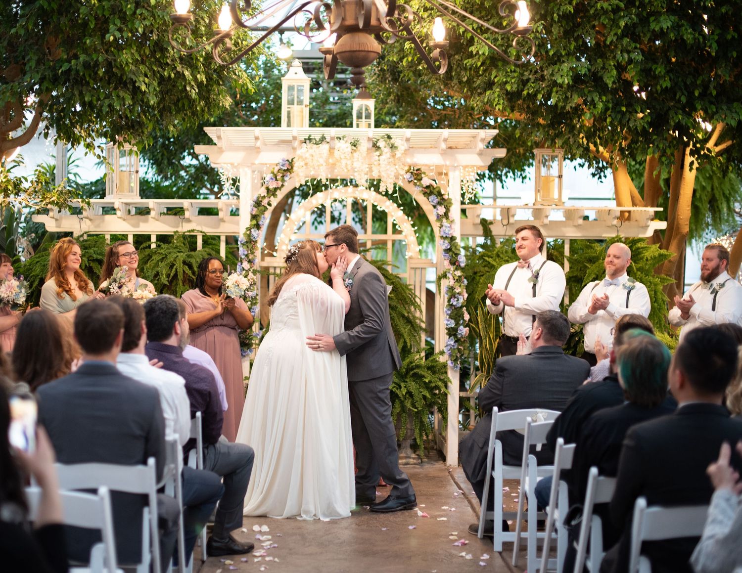 Outdoor wedding ceremony setup with white chairs and floral arch under trees.