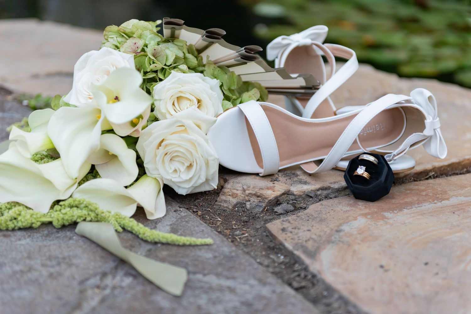 White bridal heels, wedding bouquet with roses and calla lilies, and a ring box arranged on stone pavers.