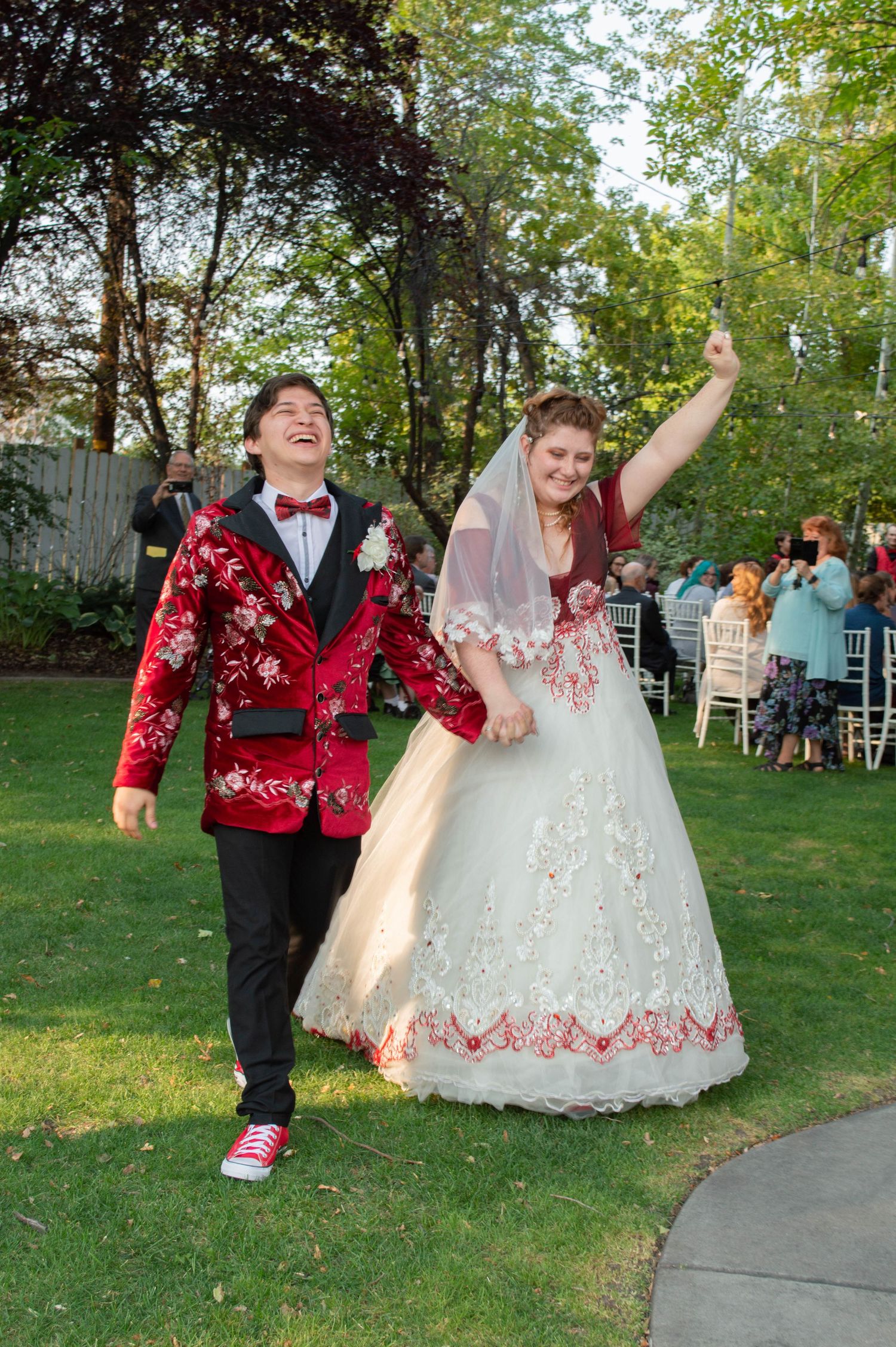 A couple in red and white formal attire dance joyfully on an outdoor lawn during a celebration.