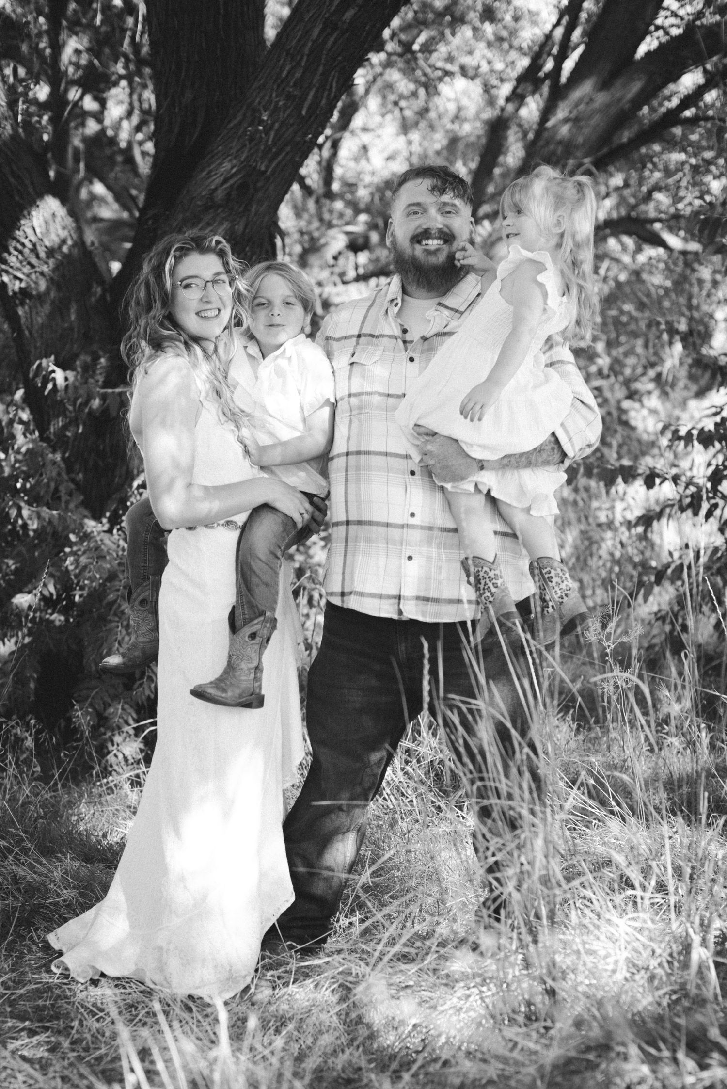 Black and white family portrait of smiling couple holding two young children outdoors among trees and tall grass.