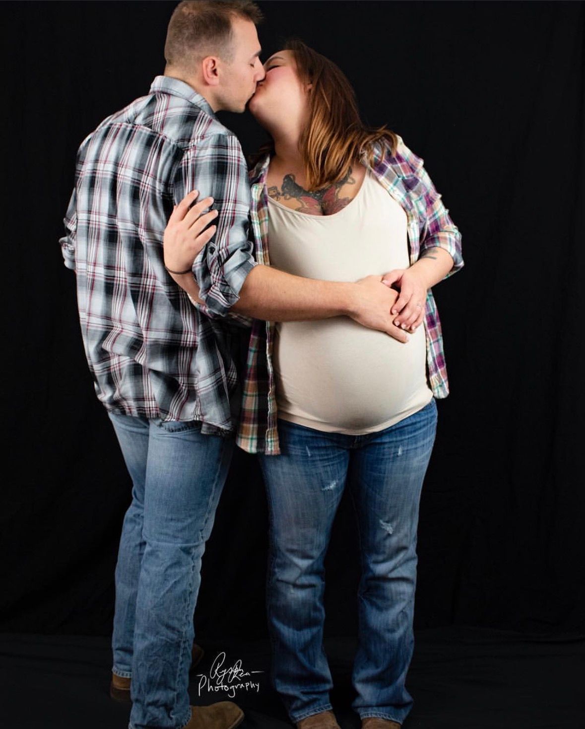 A couple in plaid shirts and jeans embrace tenderly against a black background during a maternity photoshoot.