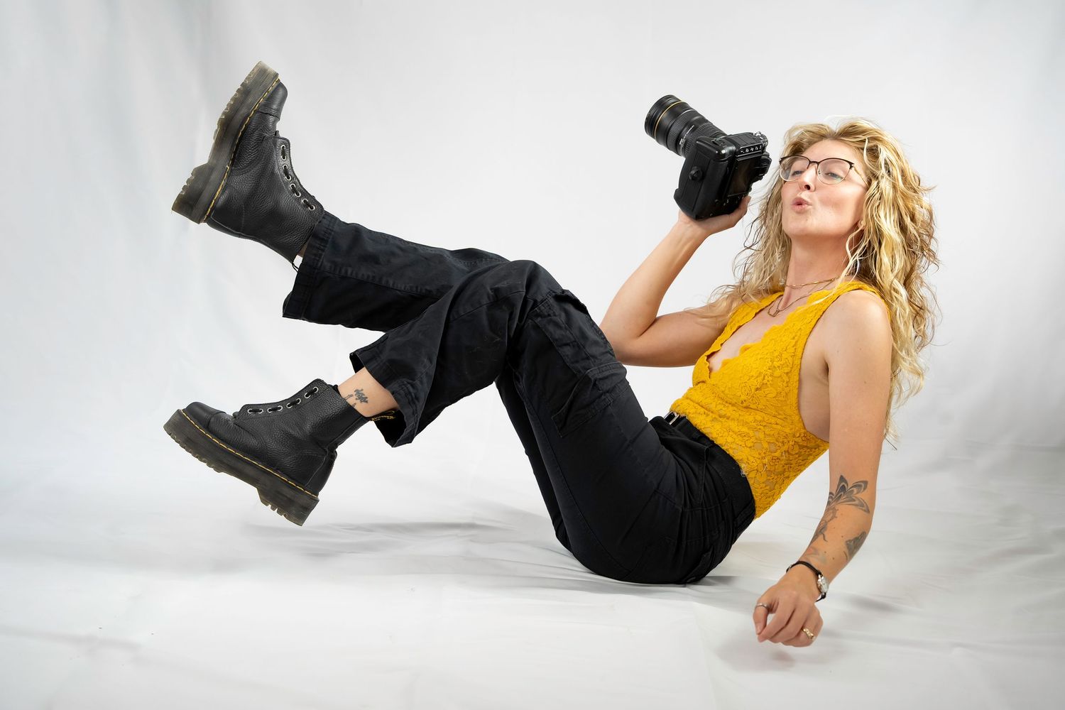 Blonde woman in yellow top and black jeans lying on floor holding camera, wearing Dr. Martens boots.