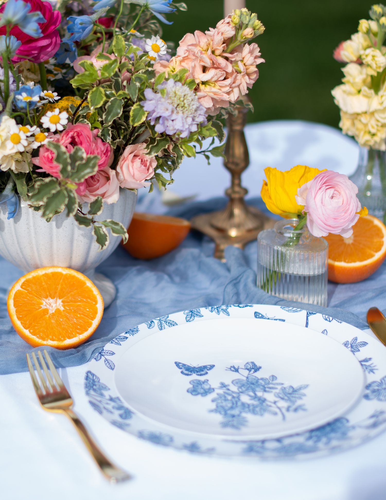 Blue and white floral china plates with fresh oranges and colorful flower arrangements on a light blue tablecloth.