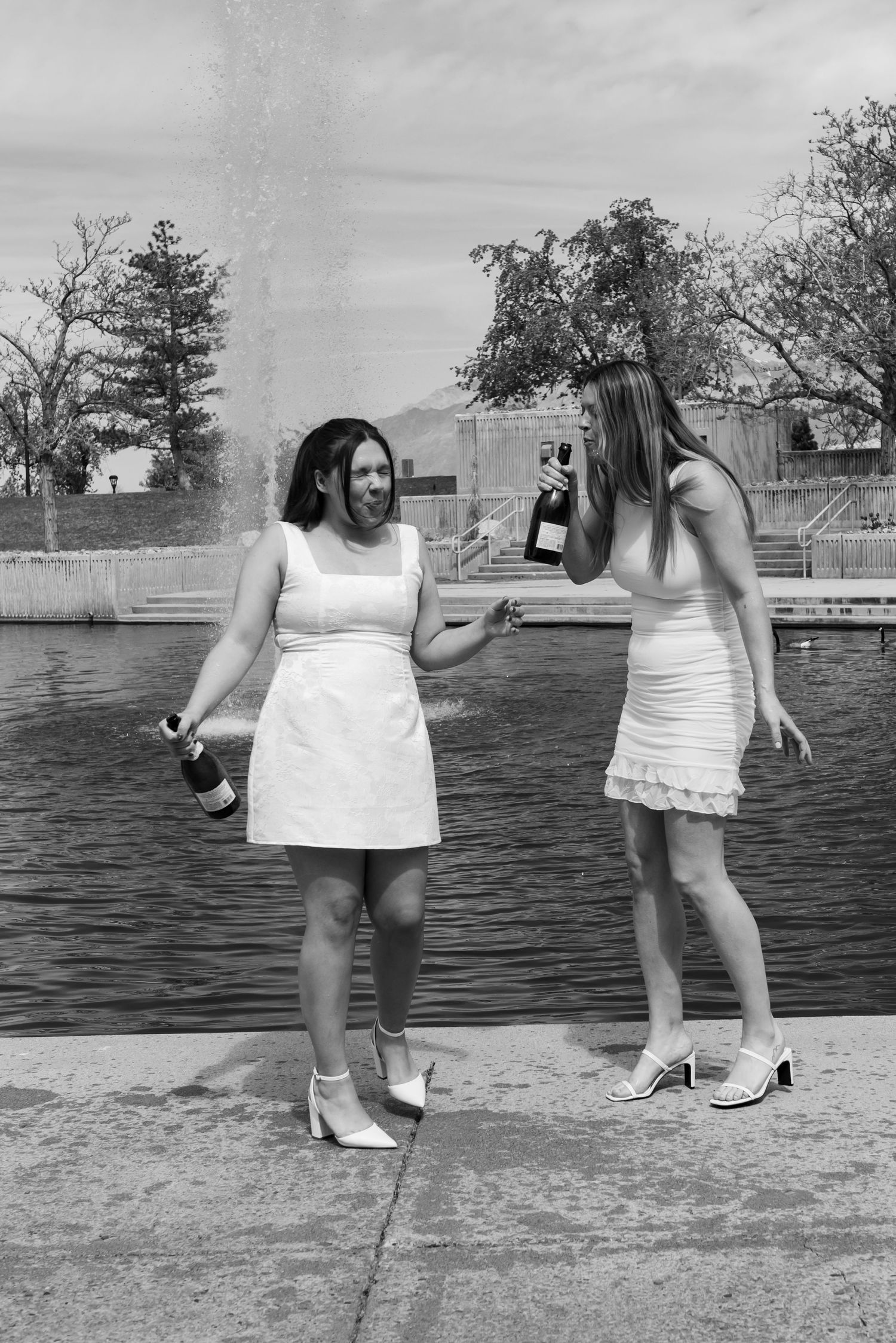 Two women in white dresses holding champagne bottles by a fountain, black and white photo.