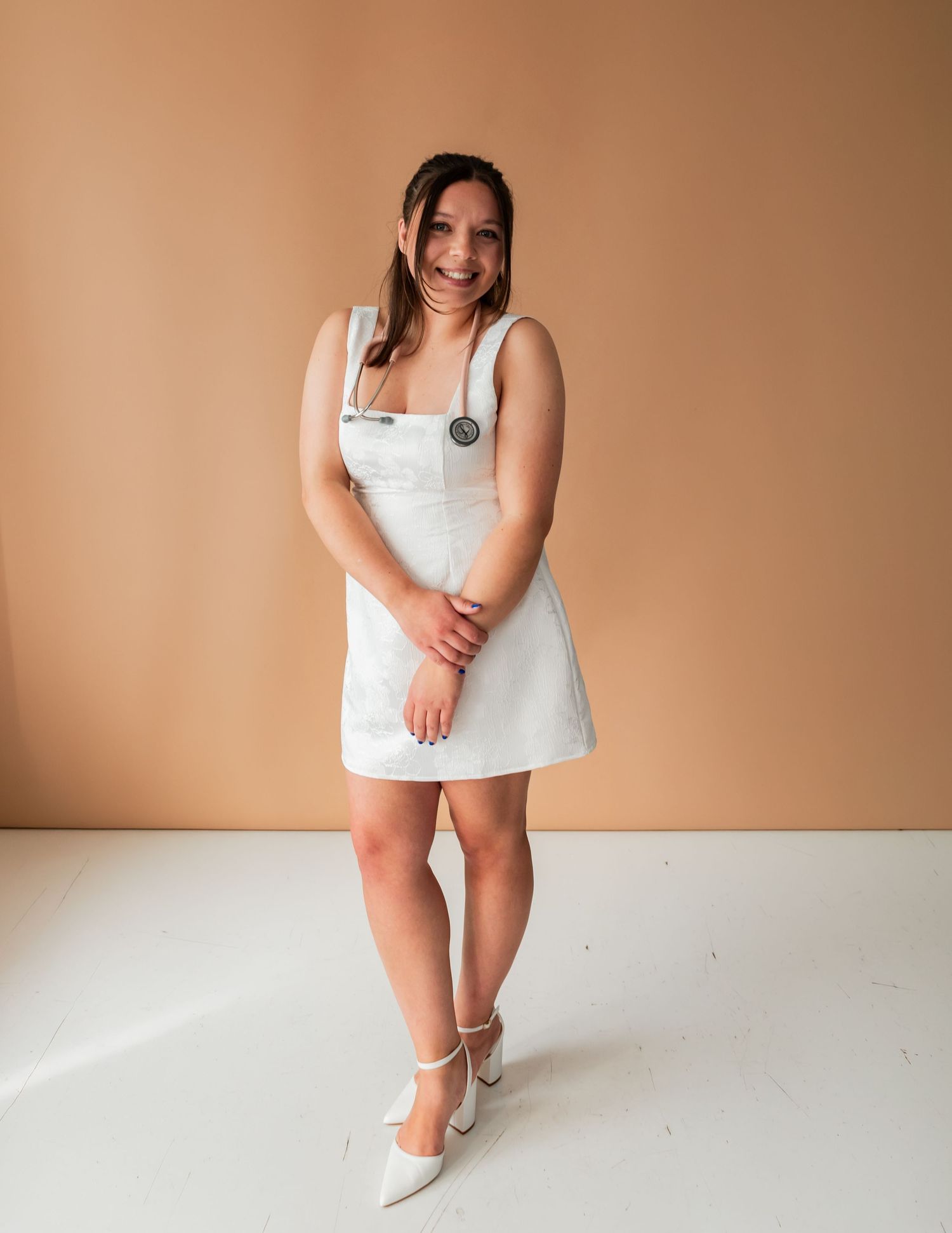 Woman in white mini dress and heels smiling against a tan background.