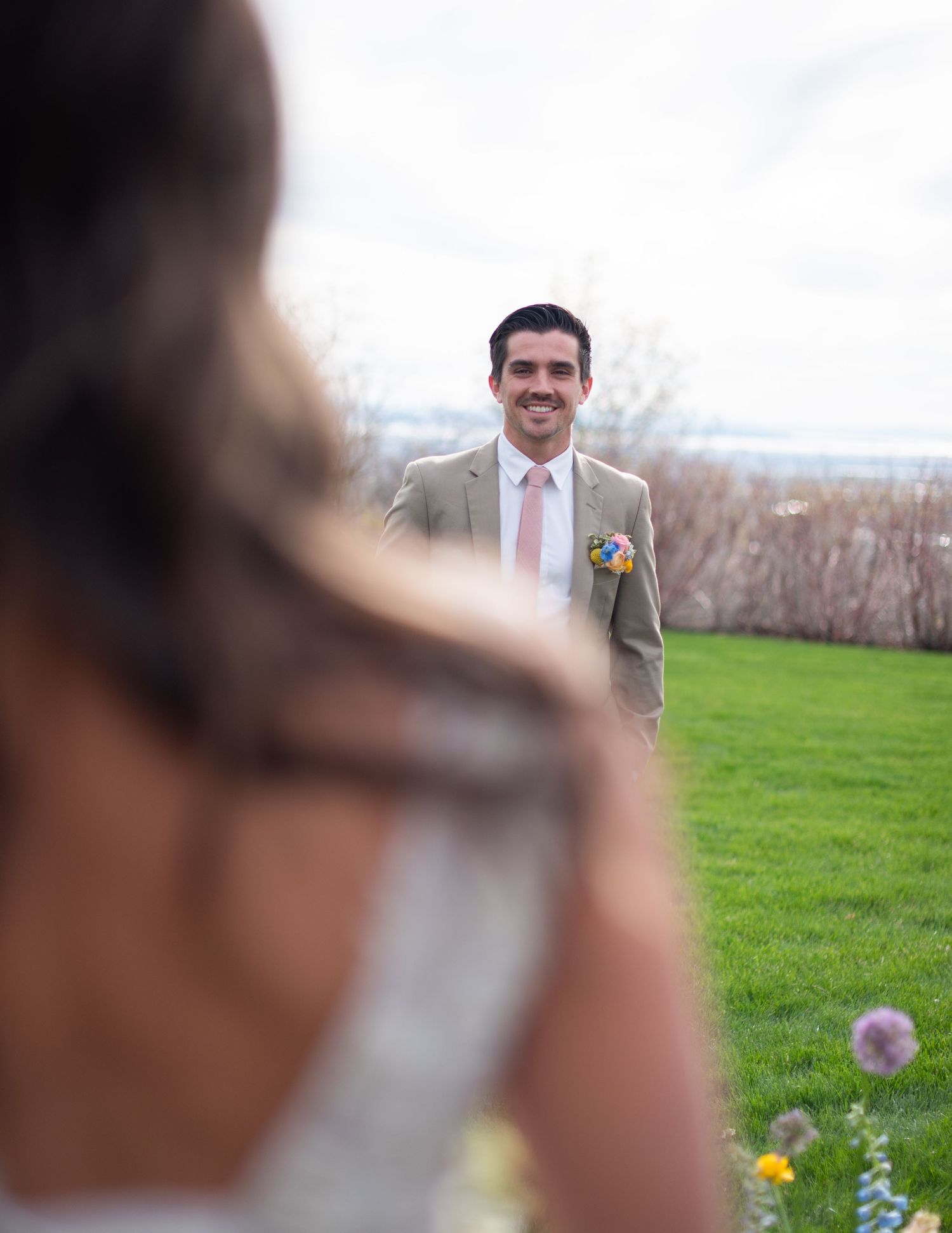 Groom in tan suit smiling at bride during outdoor wedding first look on green lawn.