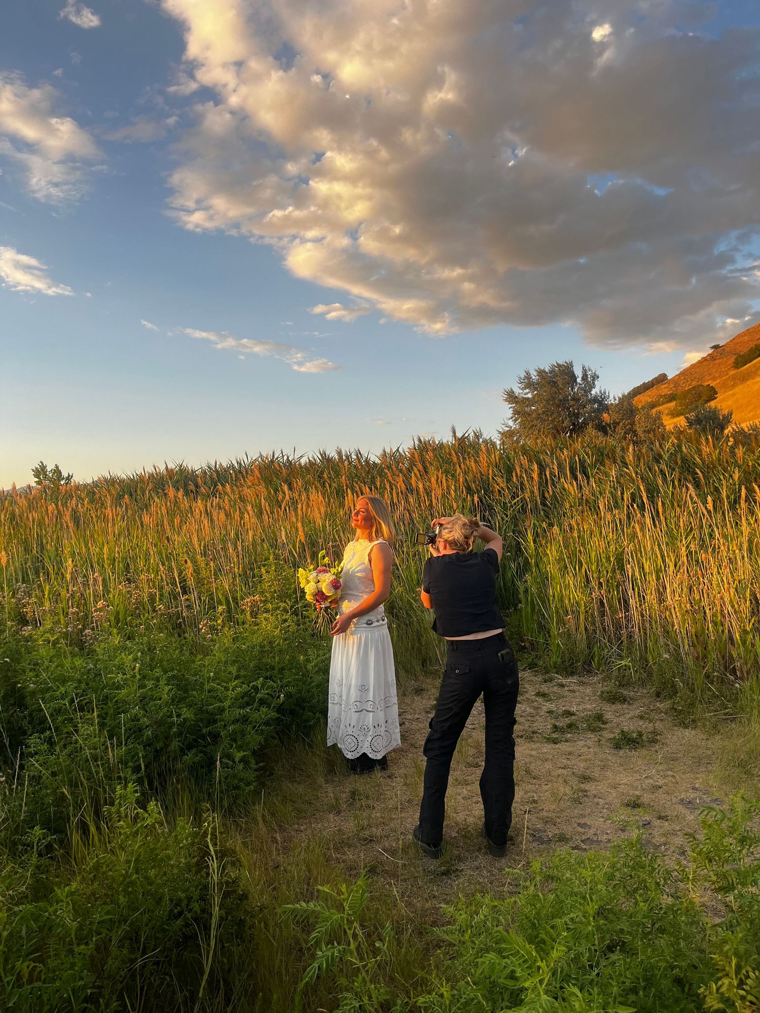 Couple walking through tall cornfield at golden hour with dramatic clouds overhead.