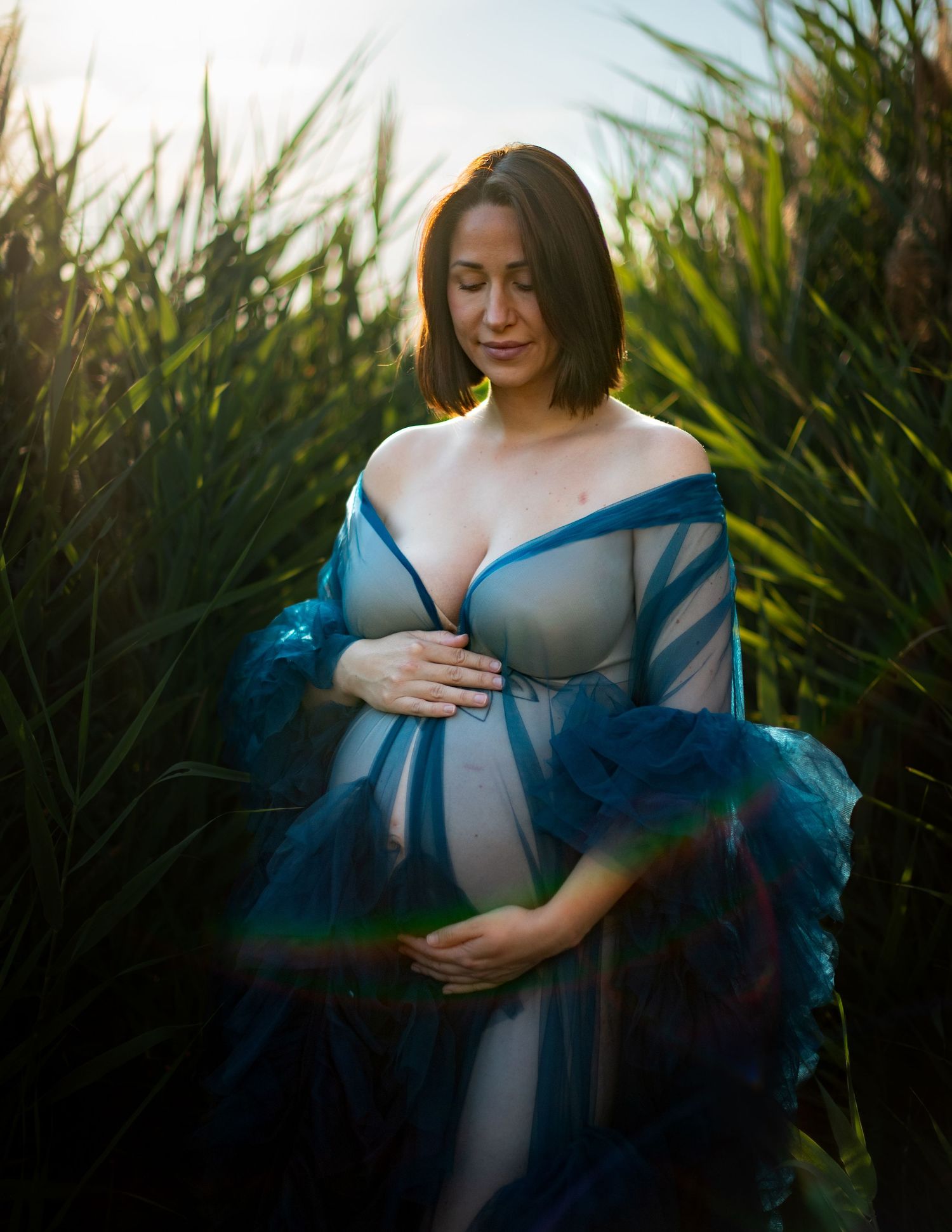 Maternity photoshoot in tall grass at sunset with flowing blue dress and natural lighting.