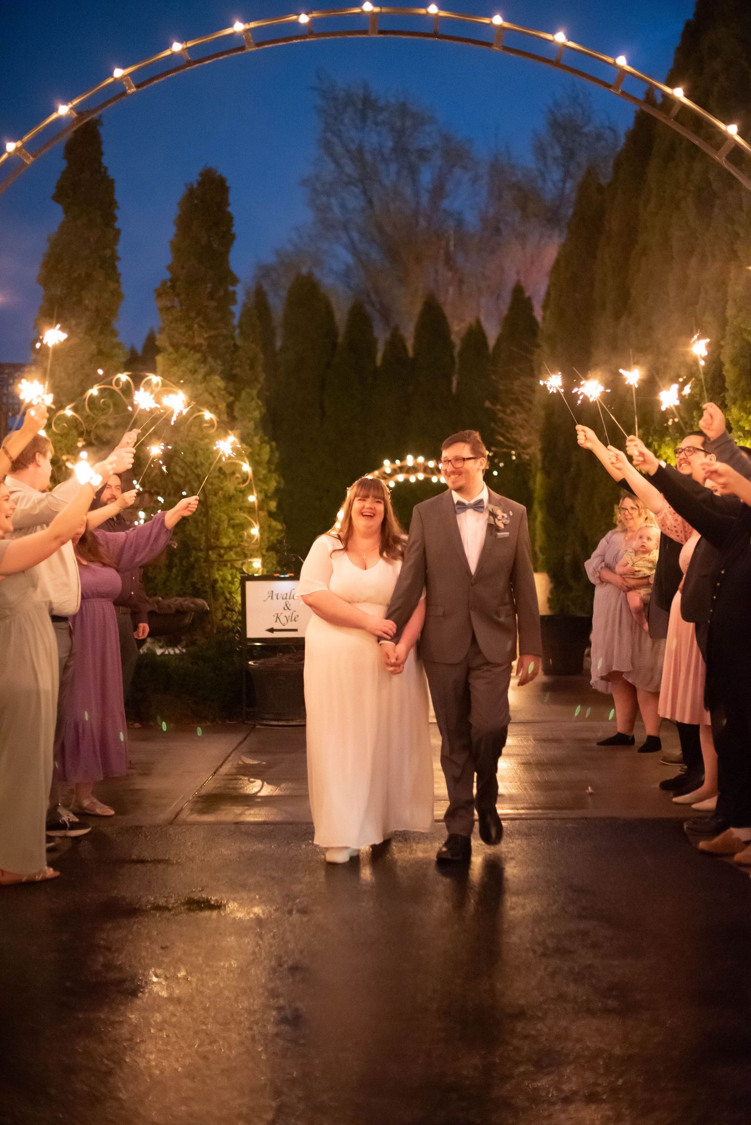 Wedding party exits under an illuminated archway at night with sparklers.