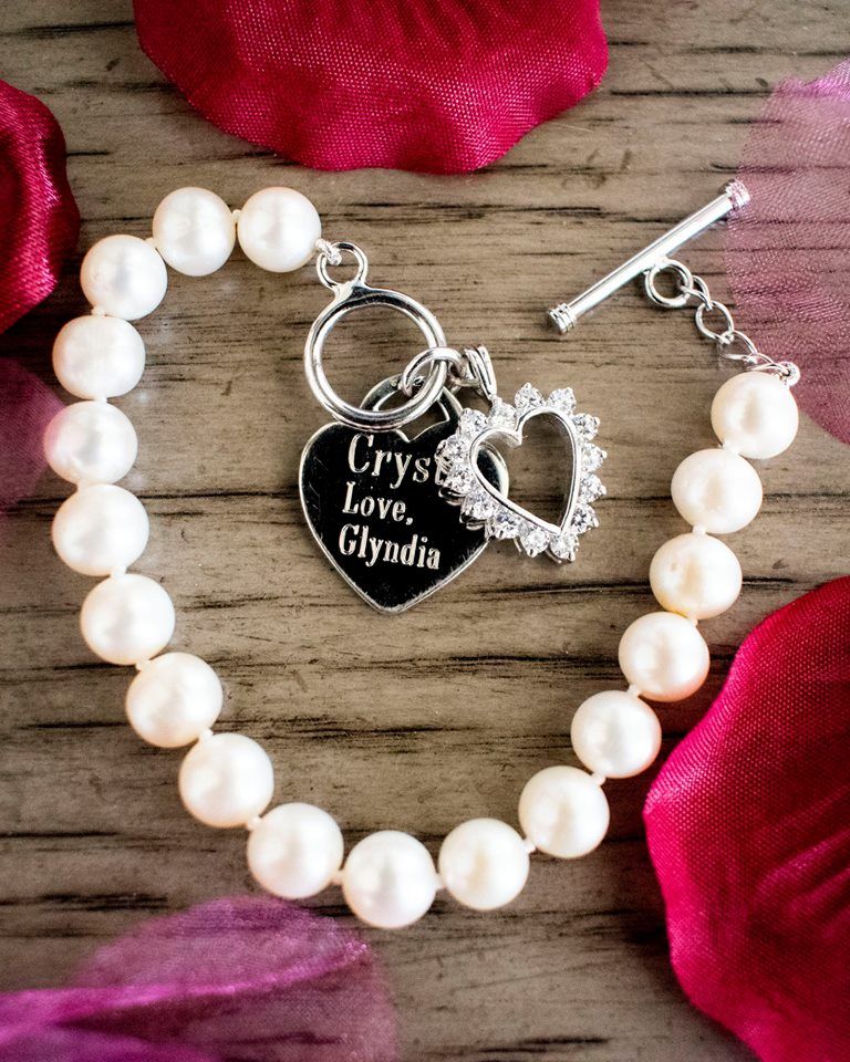 Pearl bracelet with heart charm and personalized tag on rustic wood with red rose petals.