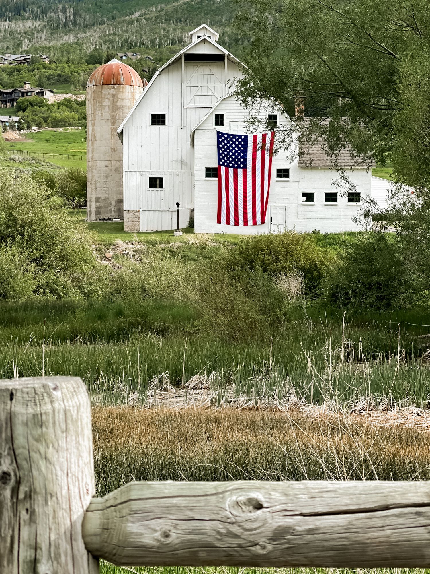White barn with American flag draped on front, silo, and rustic wooden fence in a lush green rural landscape.