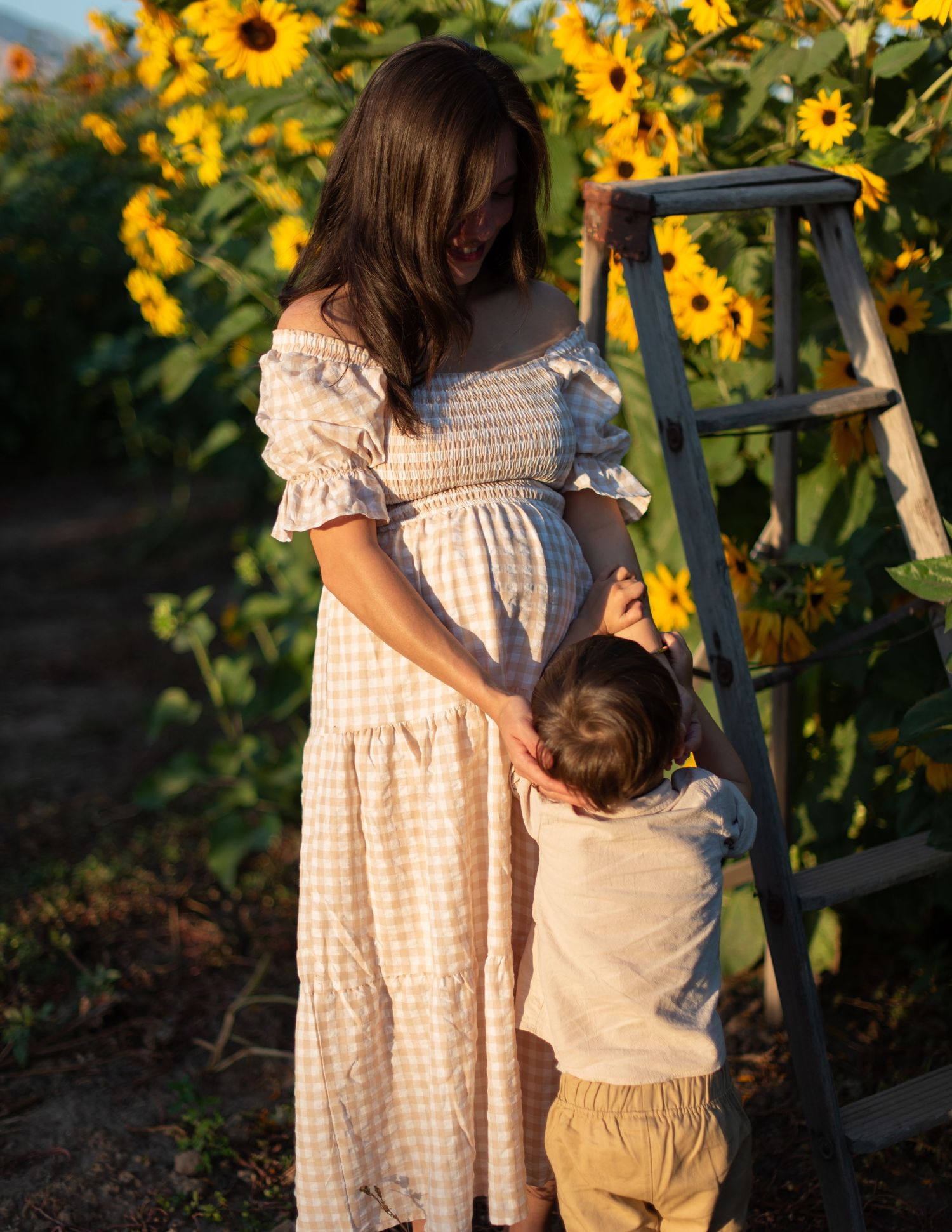 Mother and child share tender moment in sunflower field wearing matching gingham dresses.