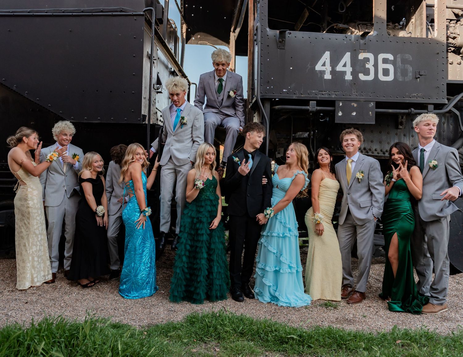 A large group of teens in formal prom attire pose together in front of a vintage train numbered 4436.