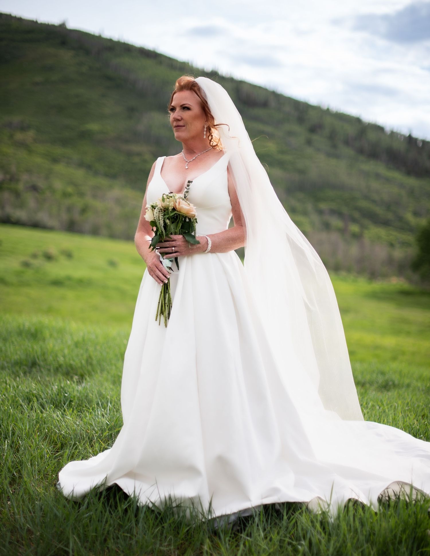 Bride in flowing white wedding gown holding bouquet in mountain meadow setting.