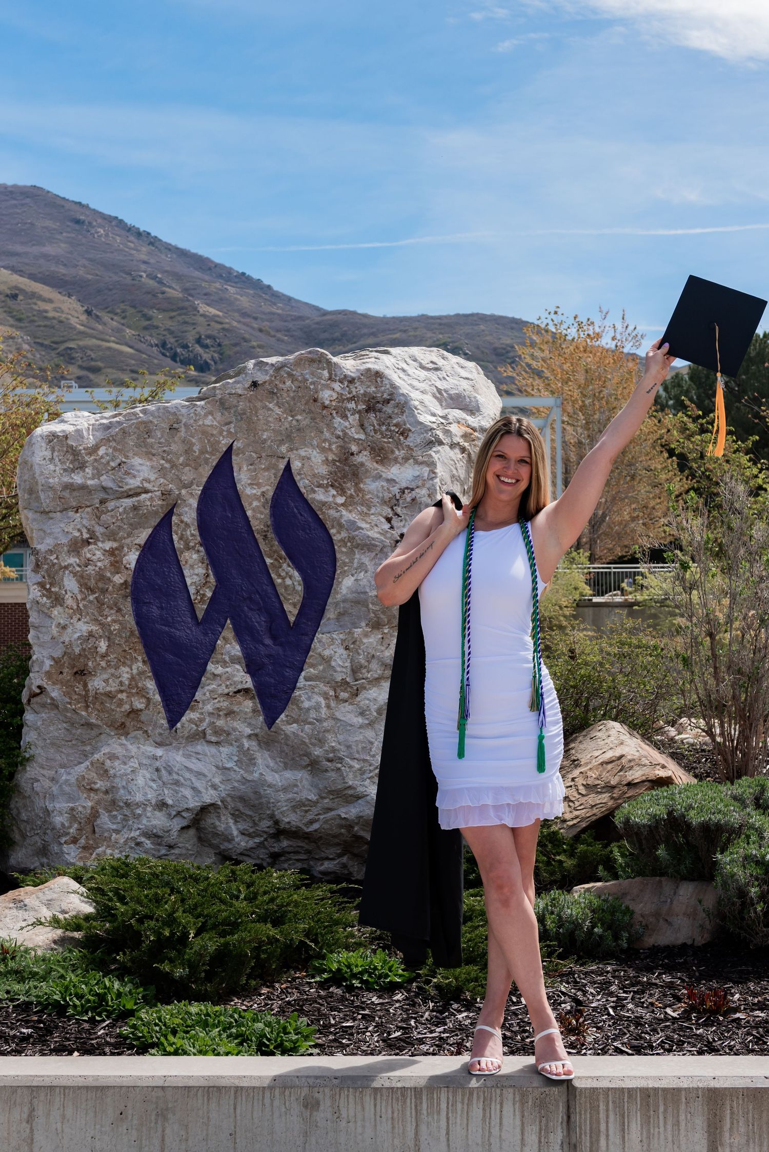 A smiling female graduate in white dress holds cap aloft beside a large rock with purple W logo, mountains behind.