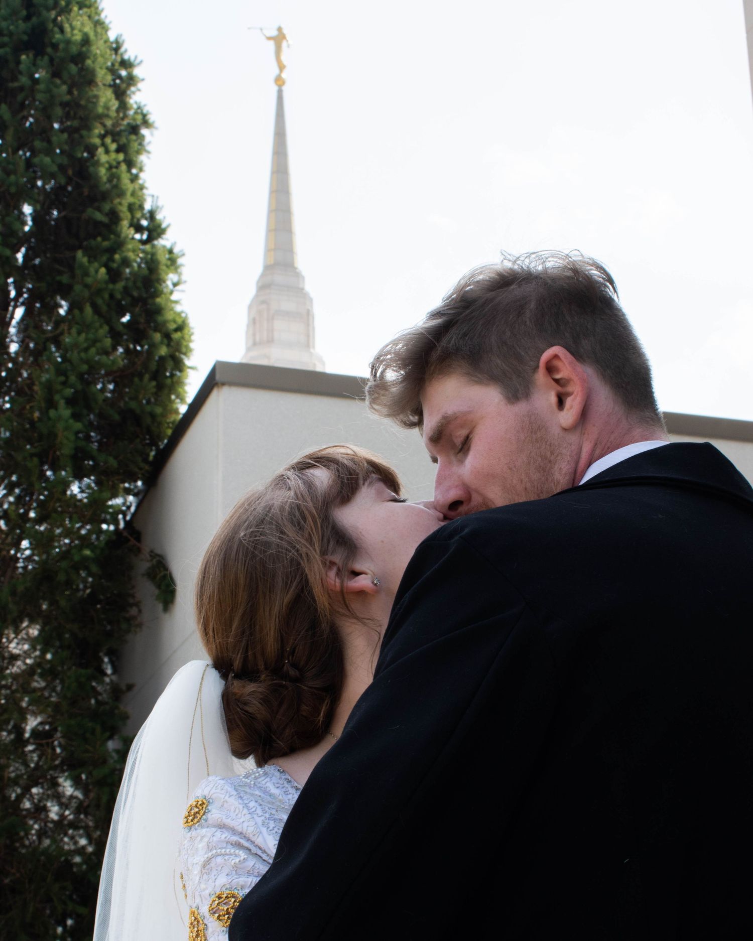 Romantic wedding couple shares a tender kiss outside a white church with a steeple in the background.