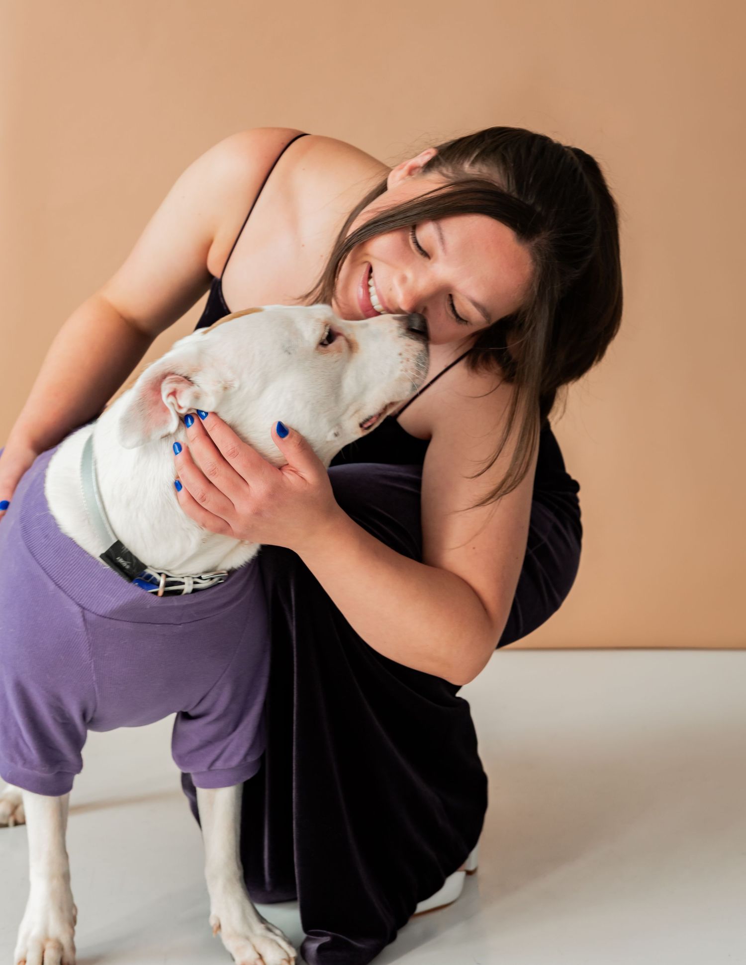 A smiling woman with dark hair hugs a white dog wearing a purple outfit against a beige background.