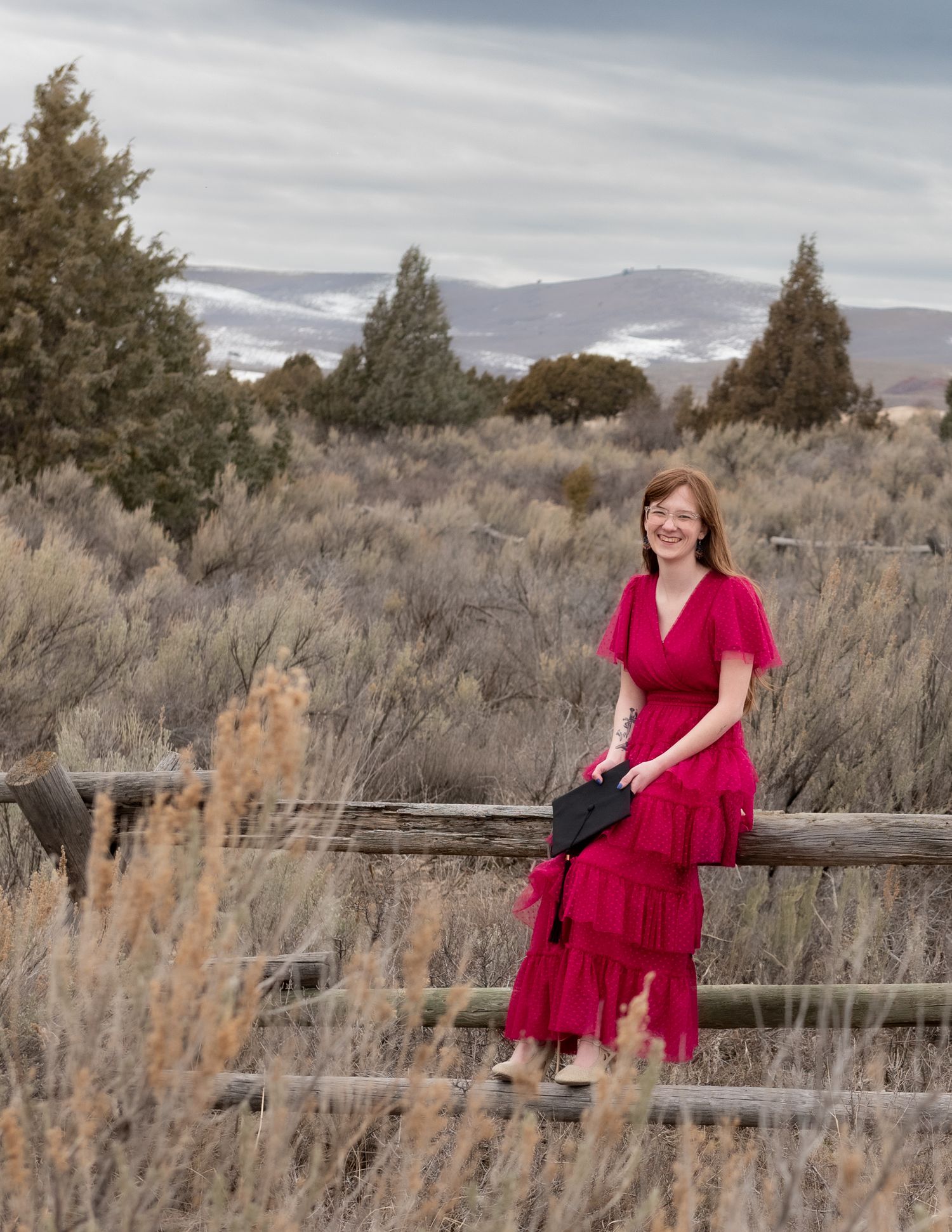 Woman in red dress poses against rustic fence with mountain landscape backdrop.
