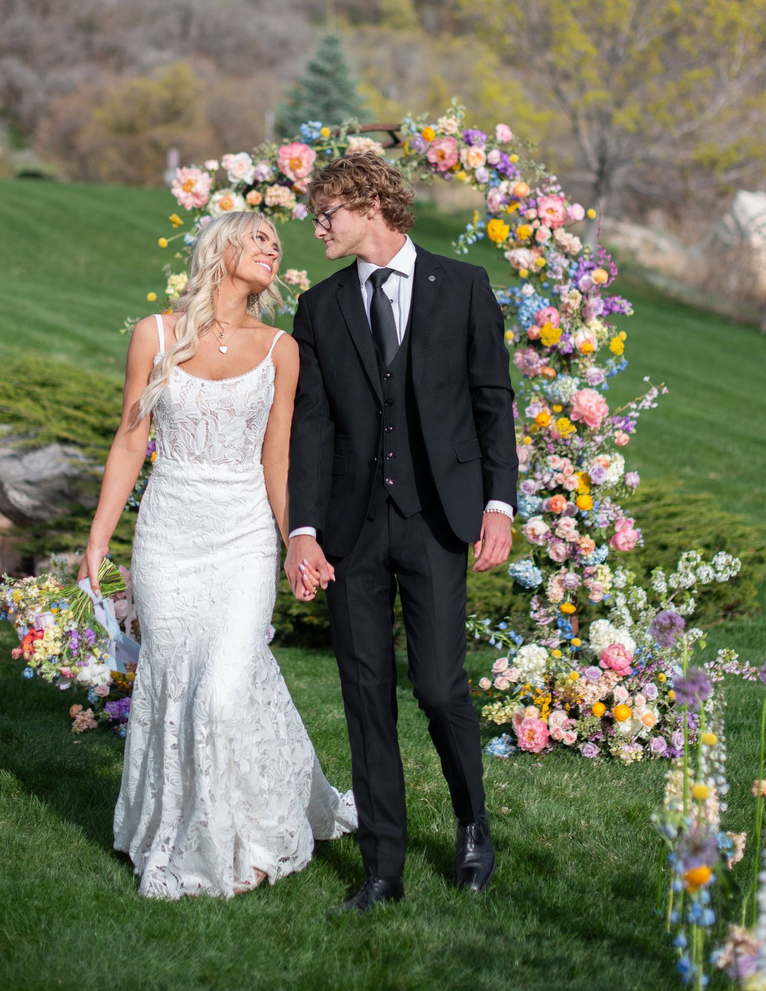 Newlyweds standing under colorful floral arch on grassy hillside.