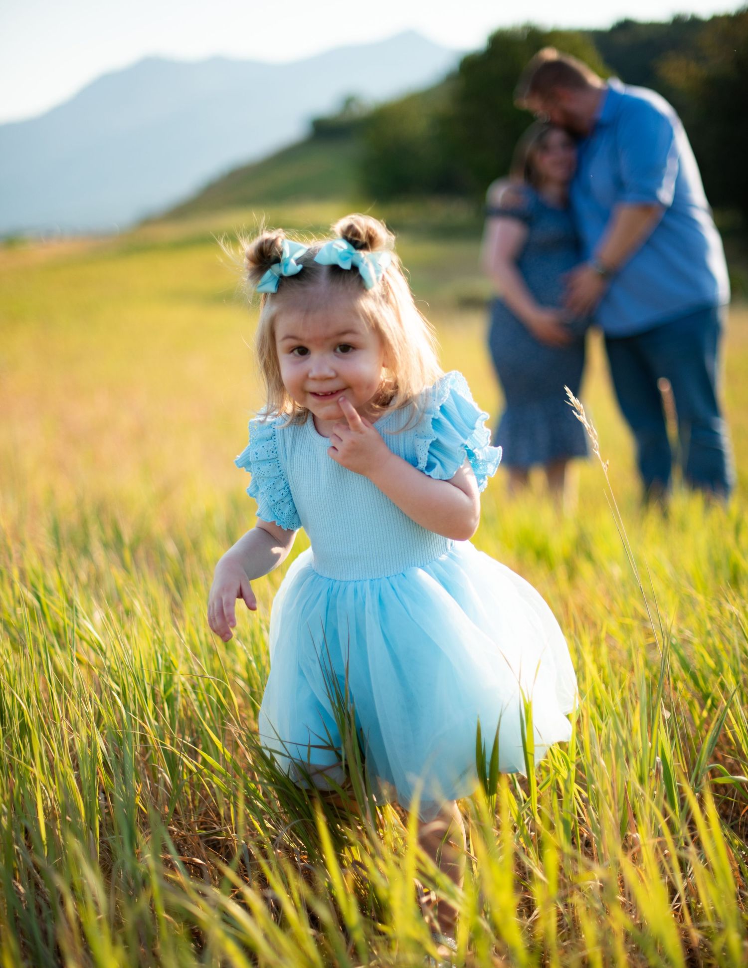 Child in blue tulle dress plays in tall grass while parents embrace in background.