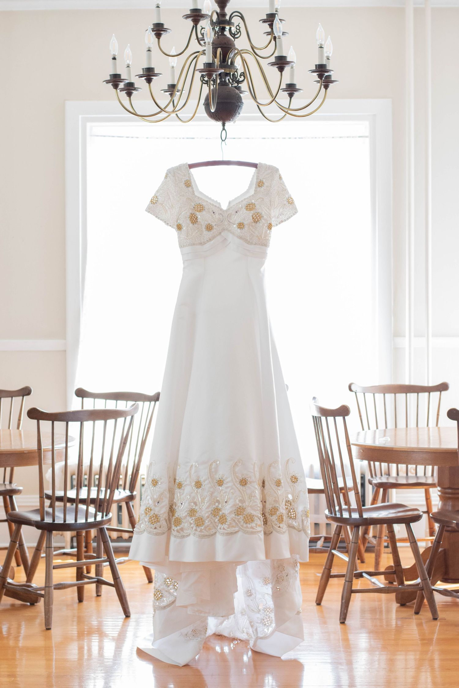 A white wedding dress with gold embellishments hangs from a chandelier in a rustic dining room.