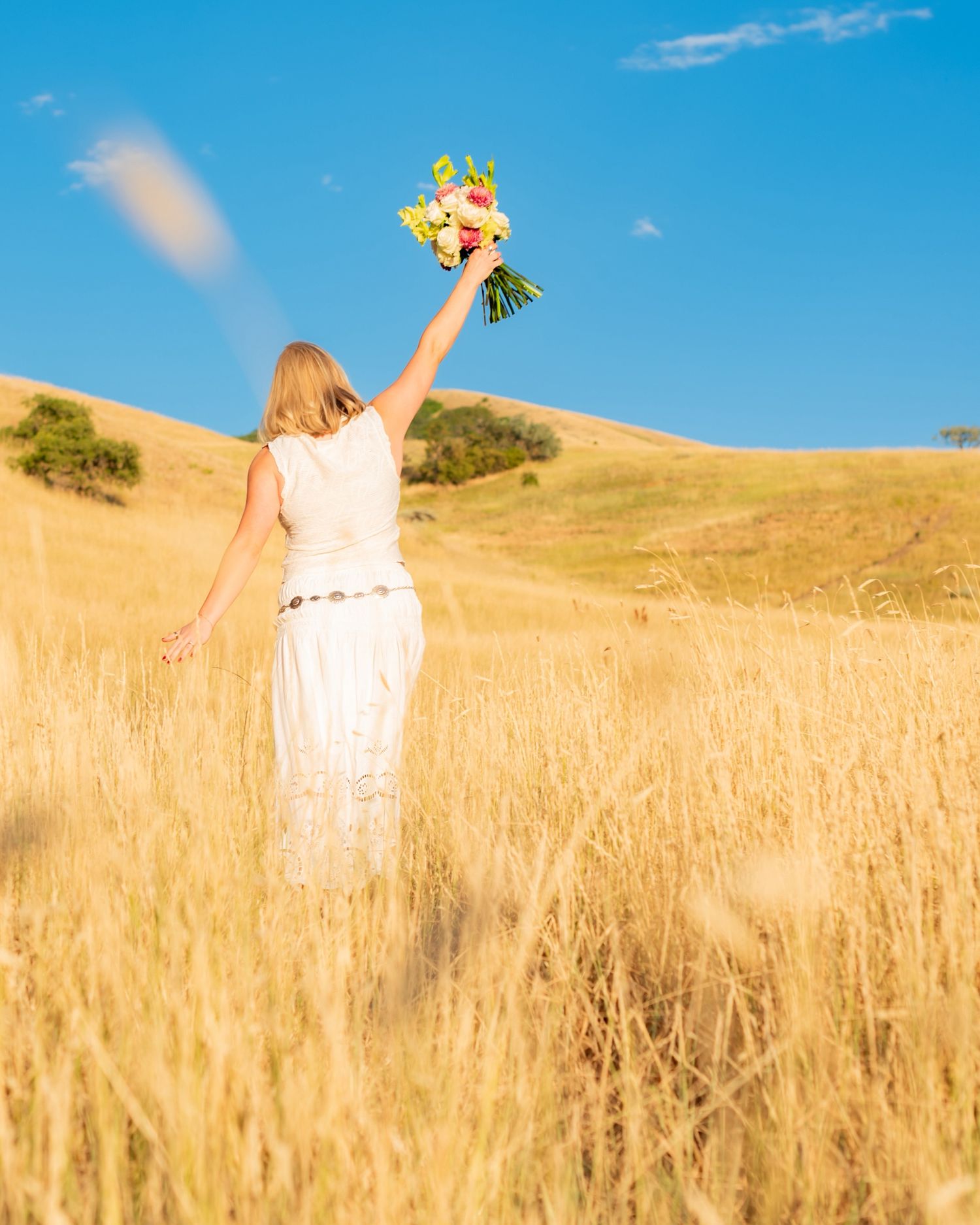 Woman in white dress holding bouquet triumphantly in golden wheat field against blue sky.