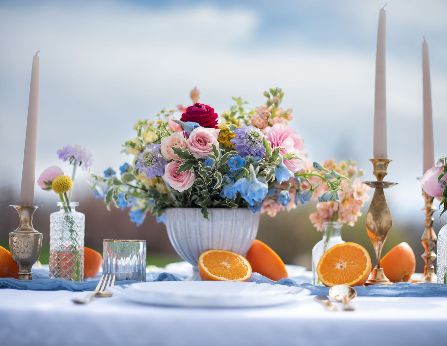 Elegant table setting with colorful floral centerpiece, candles, and fresh oranges on a blue tablecloth.