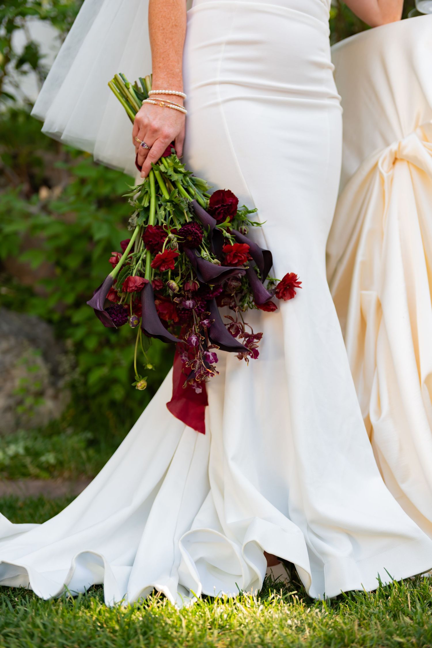 Close-up view of a white wedding dress with deep red floral bouquet against green grass.