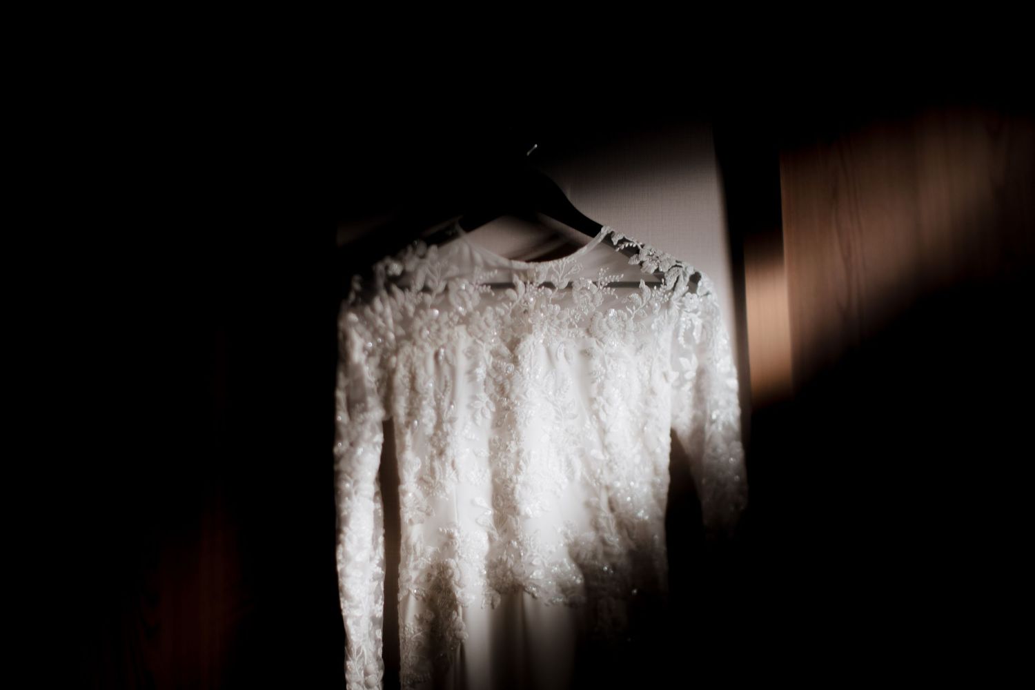 A delicate white lace wedding dress hangs in dramatic shadows with light streaming across its intricate details.