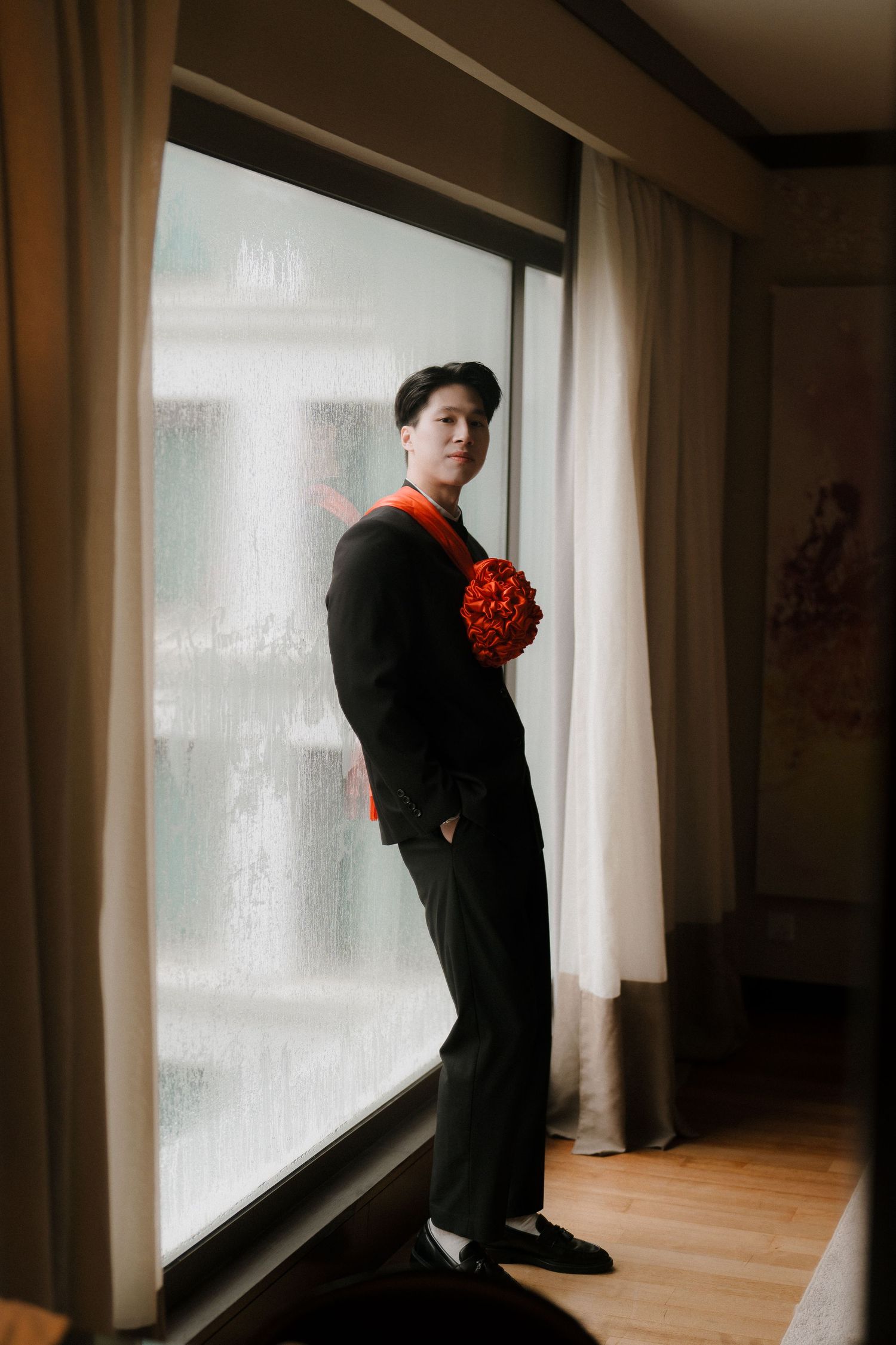 Person in formal black attire stands near window holding red bouquet captured by Crossfadefilm