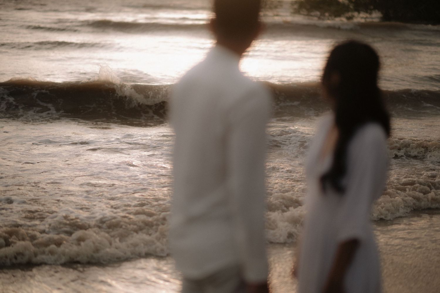 Silhouetted figures in white clothing stand apart on a sandy beach at sunset creating a moody atmosphere.