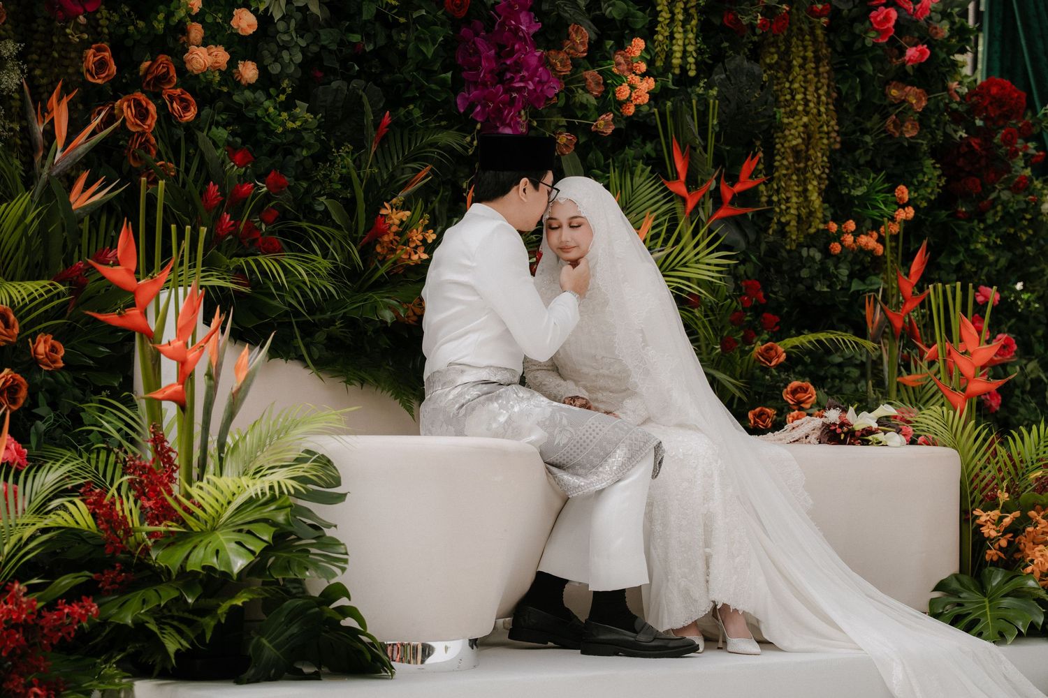 A couple in white traditional Malaysian wedding attire shares an intimate moment surrounded by lush tropical florals.