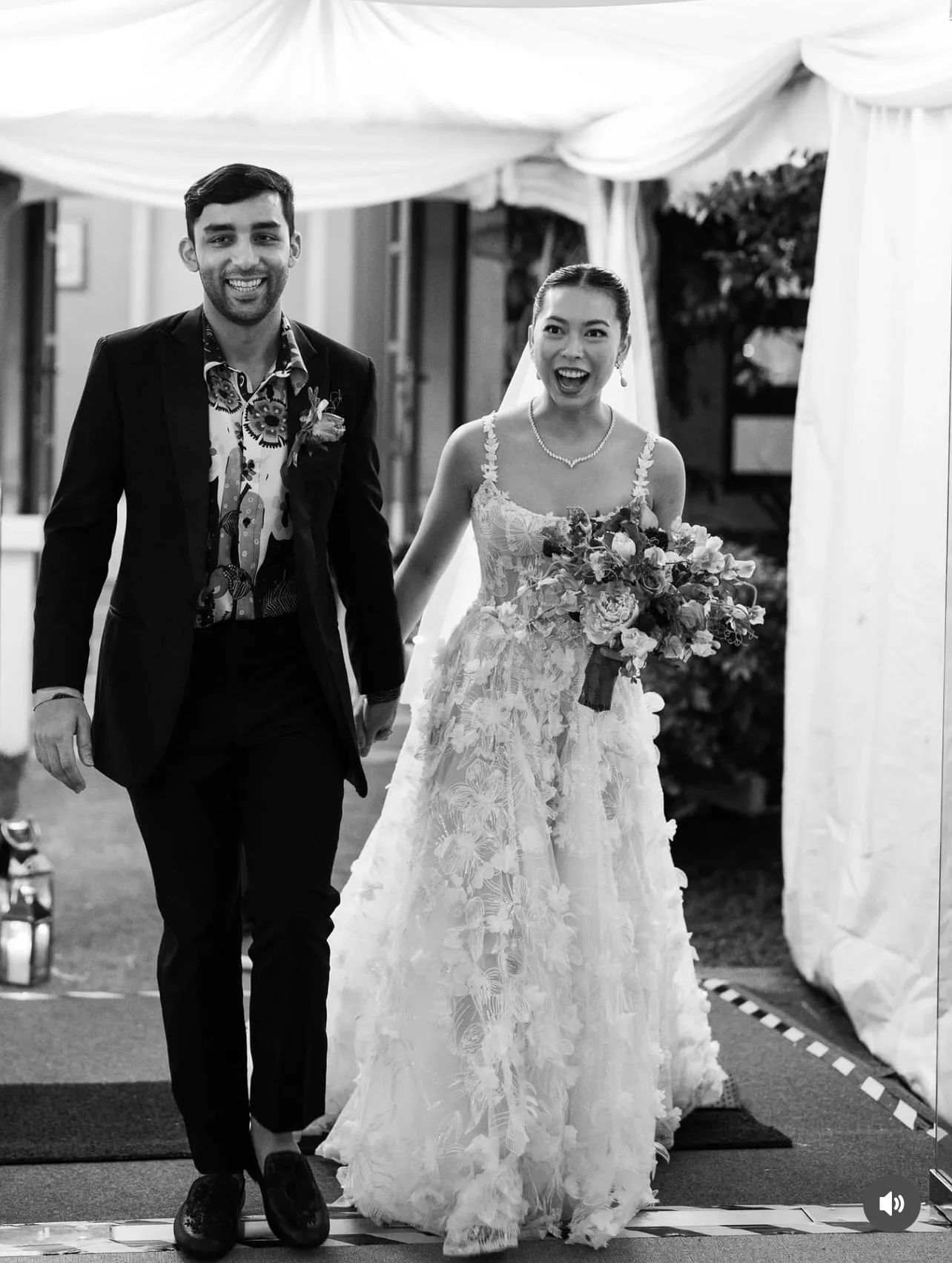 Smiling bride in a floral lace wedding gown holding a bouquet walks hand in hand with a groom in a dark suit beneath a white tent.