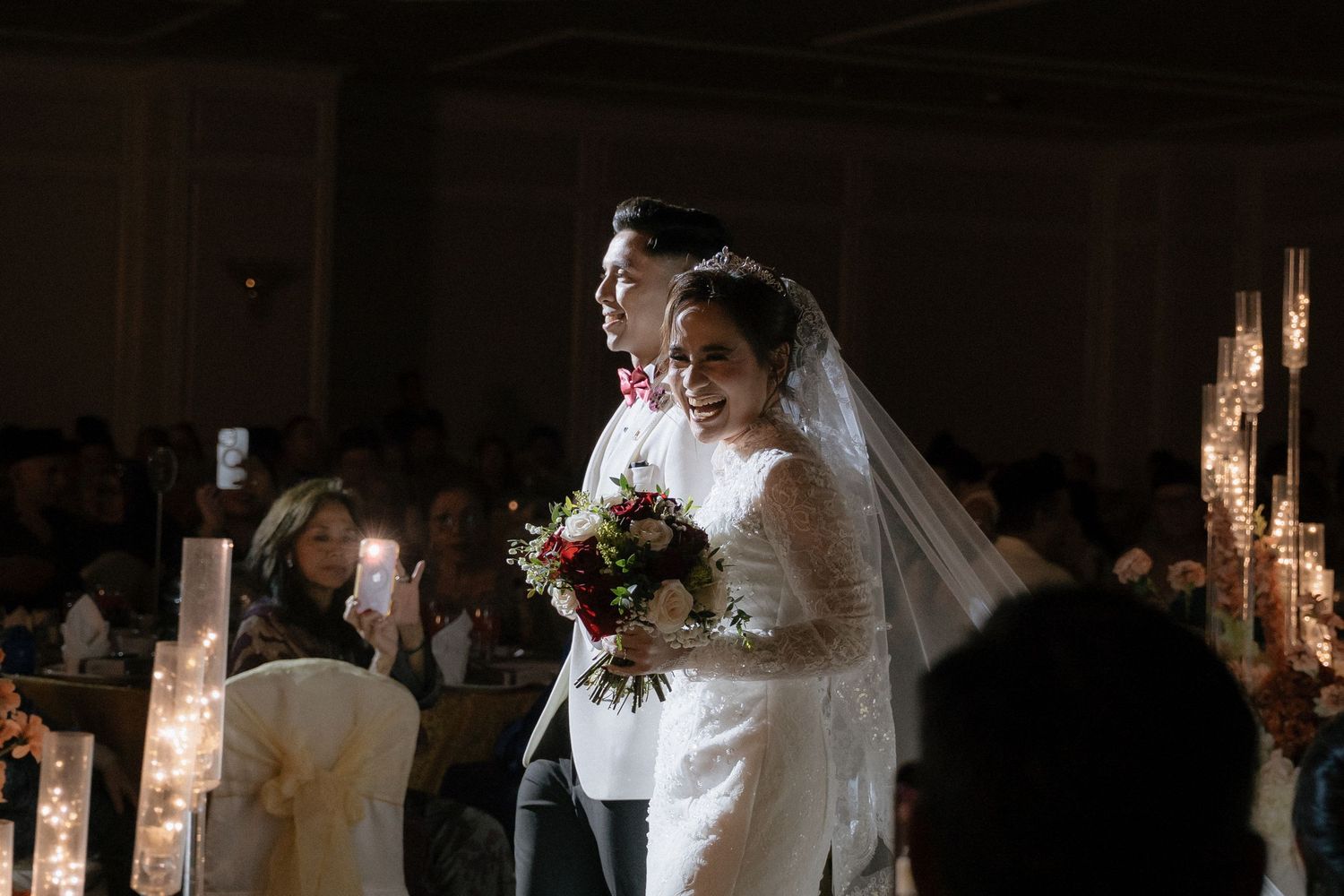 A bride and groom stand together at their wedding ceremony holding a bouquet in a dimly lit venue.