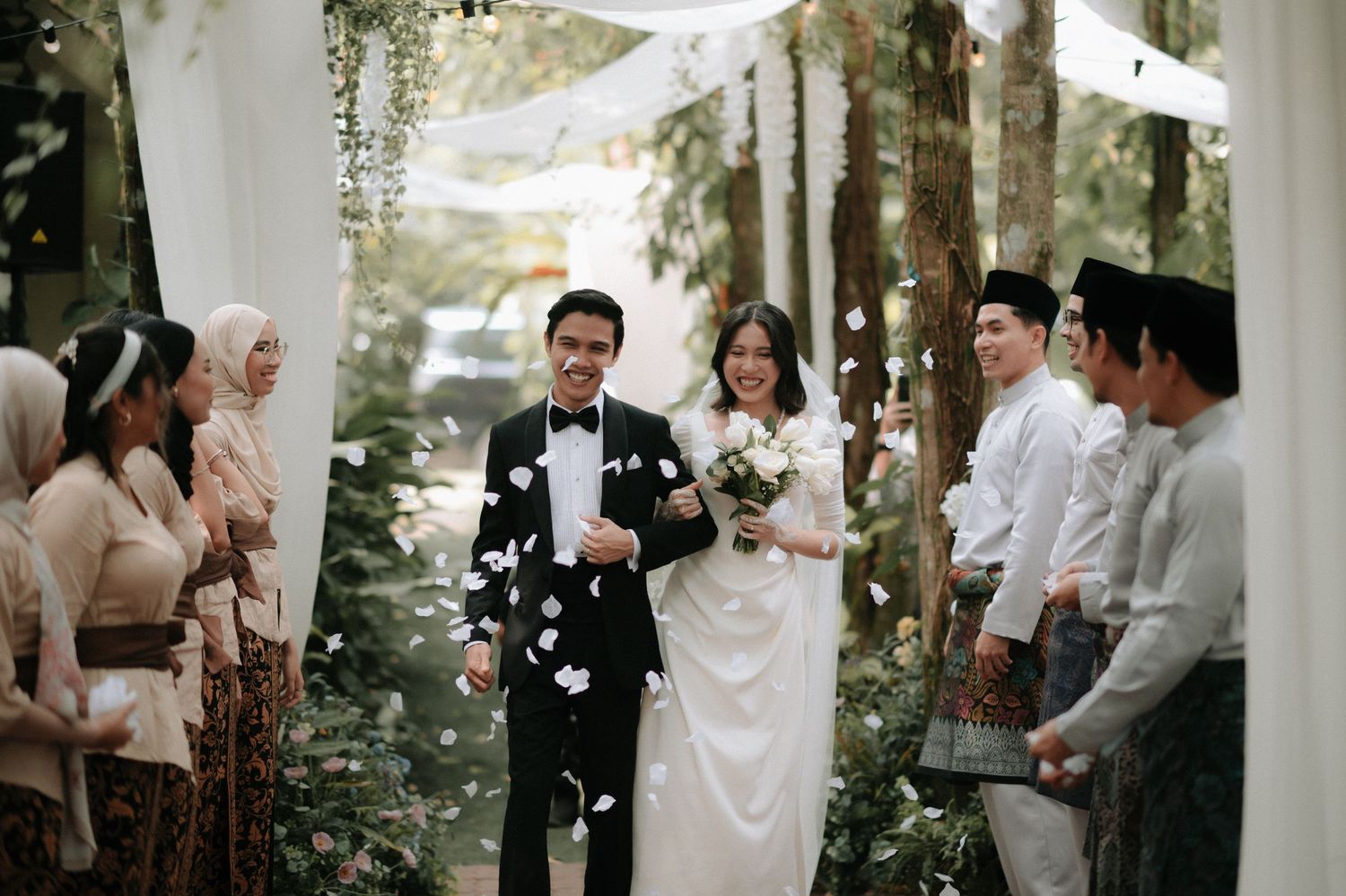 Newlyweds walk down outdoor forest aisle while guests throw white flower petals during intimate garden ceremony.