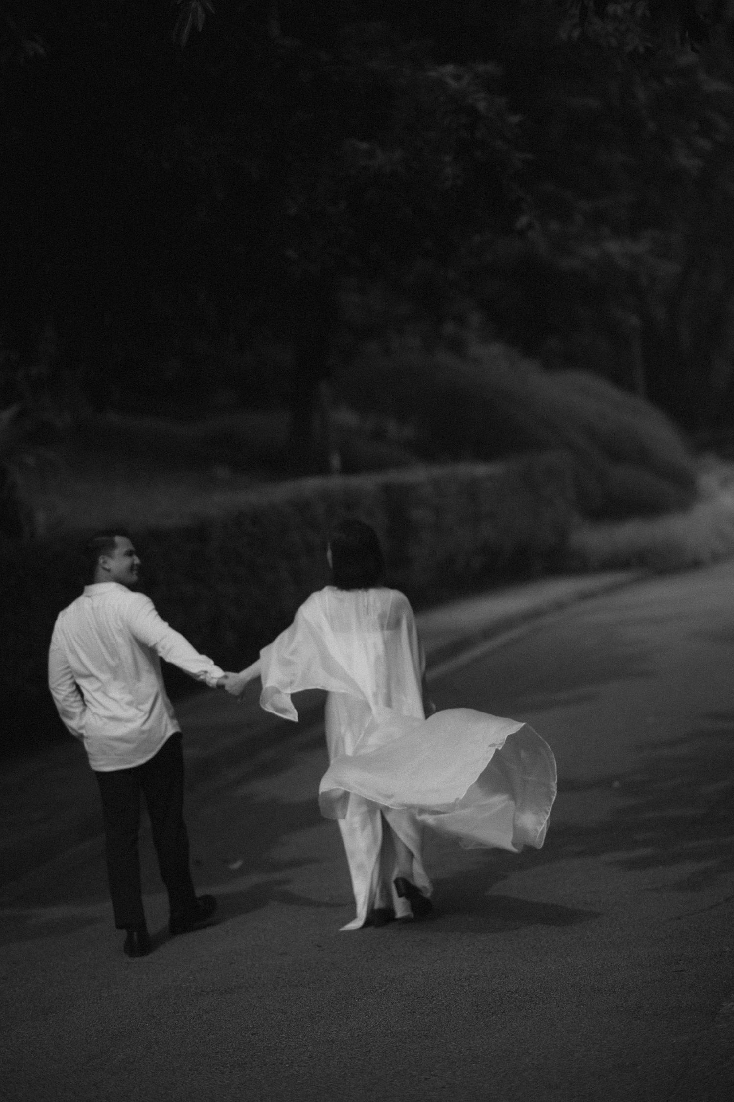 Black and white photo of a couple holding hands and walking down a tree-lined road, her flowing white gown billowing in the breeze.