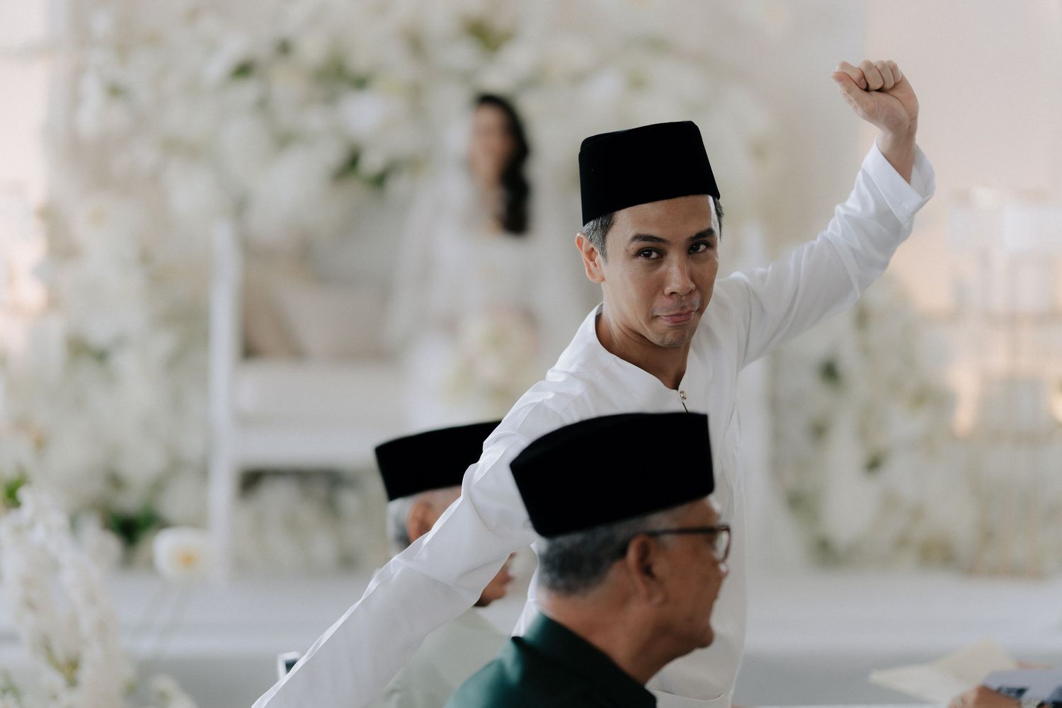 Groom in white traditional attire and black songkok raising his fist at a wedding ceremony with white floral decor and guests nearby.
